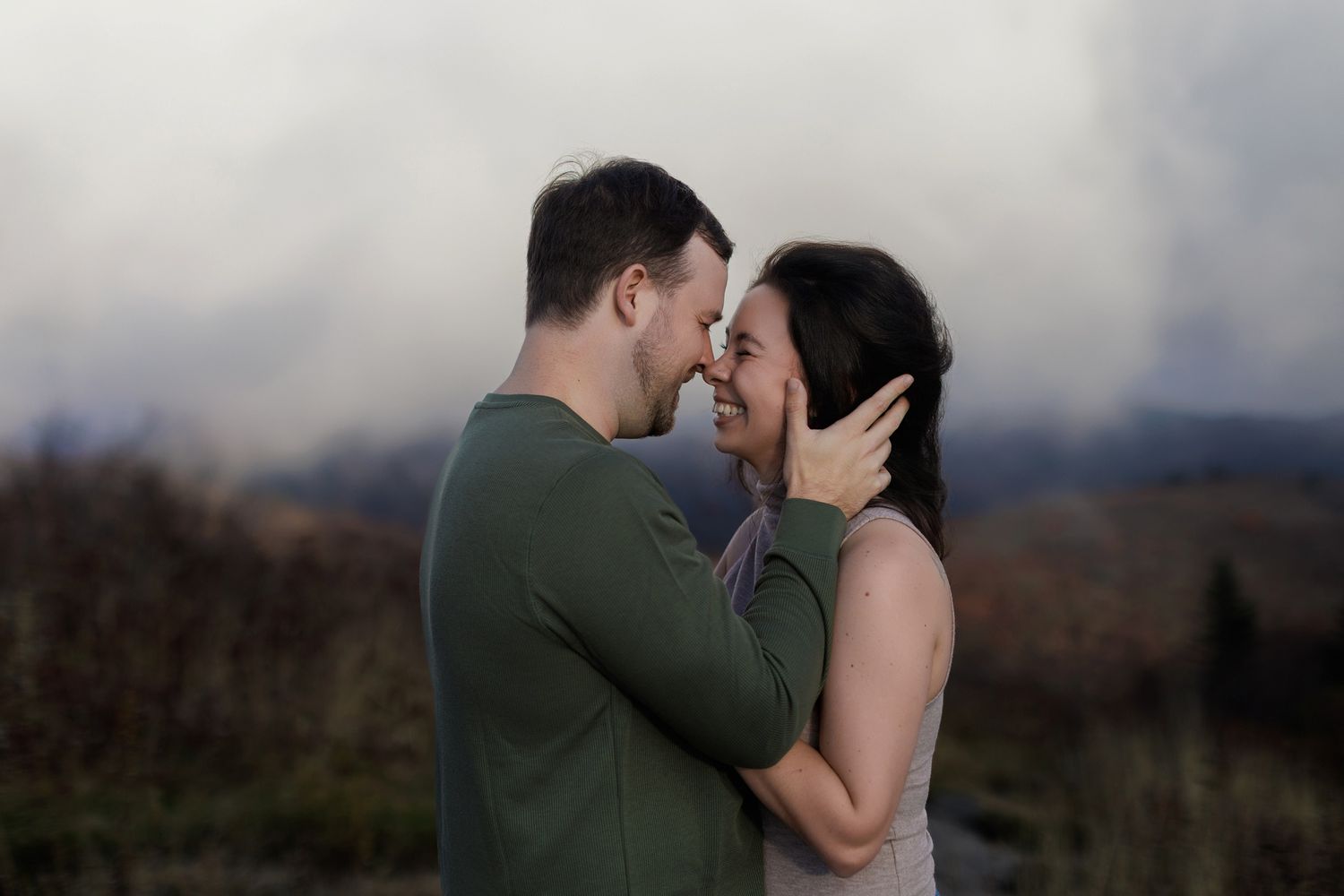 Couple embraces in romantic outdoor setting with mountains and cloudy sky in the background.