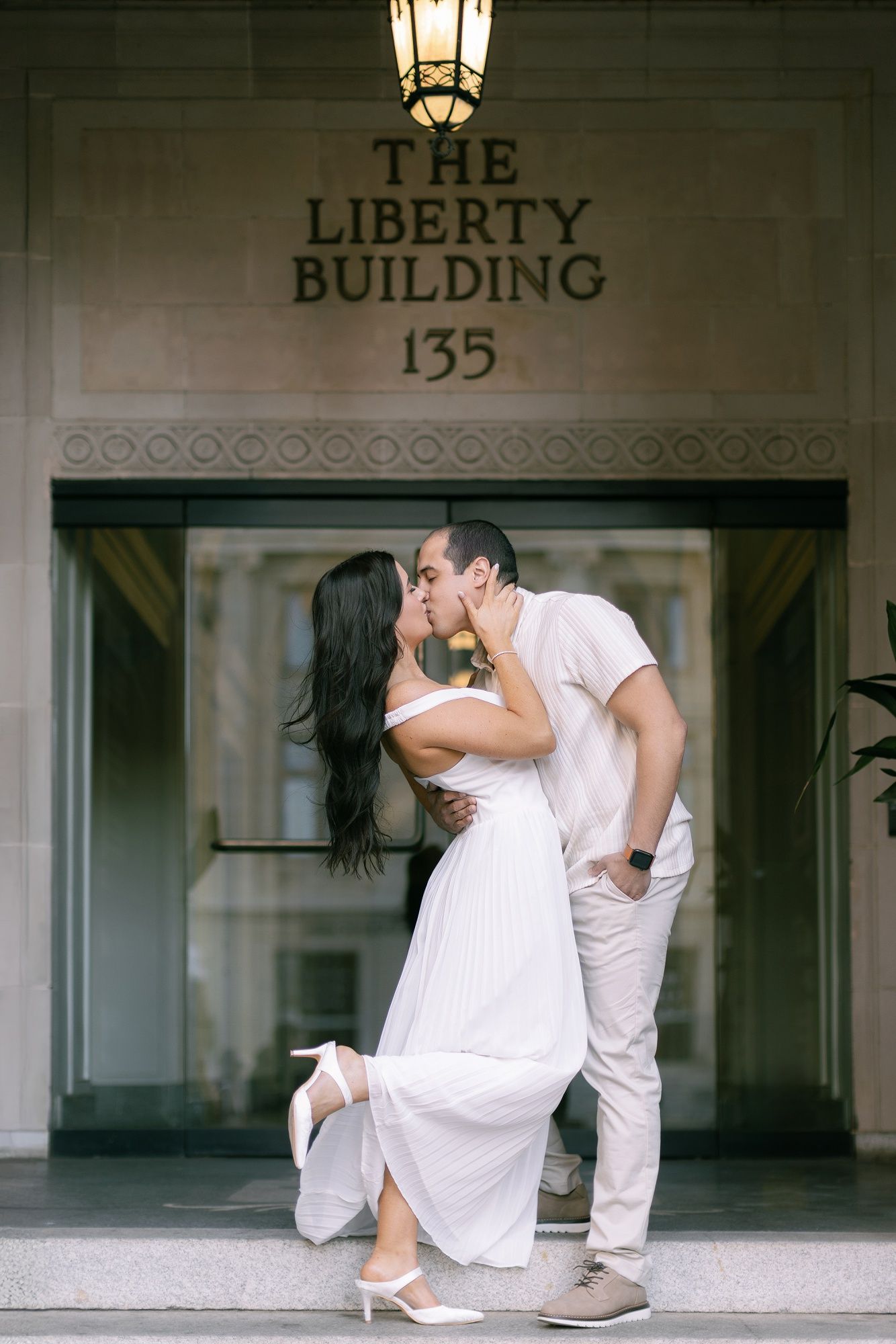 A romantic couple in white outfits share an intimate kiss in front of a building entrance with classical architecture.