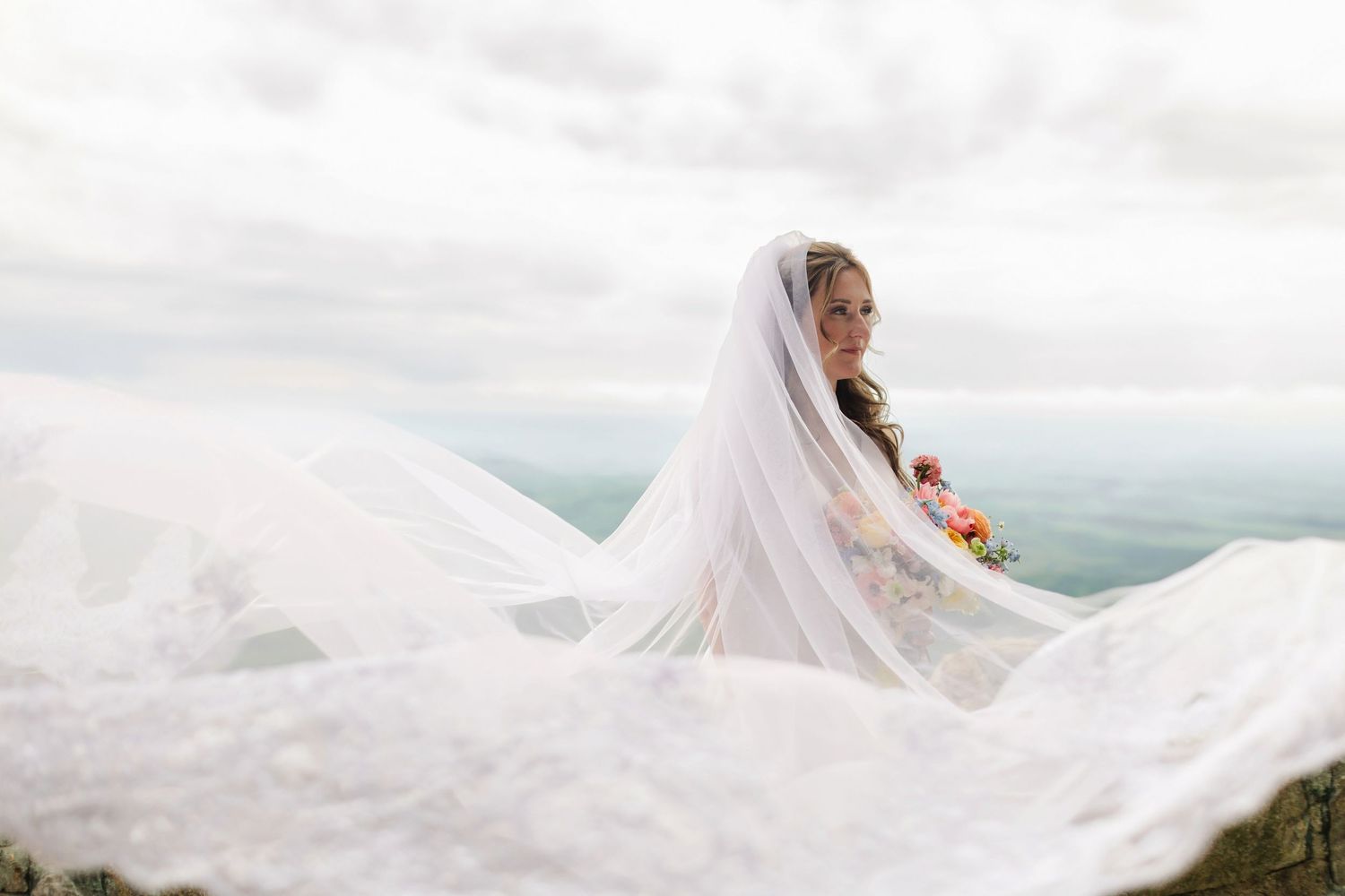 Bride in white gown with flowing veil billowing in wind against cloudy mountain backdrop.