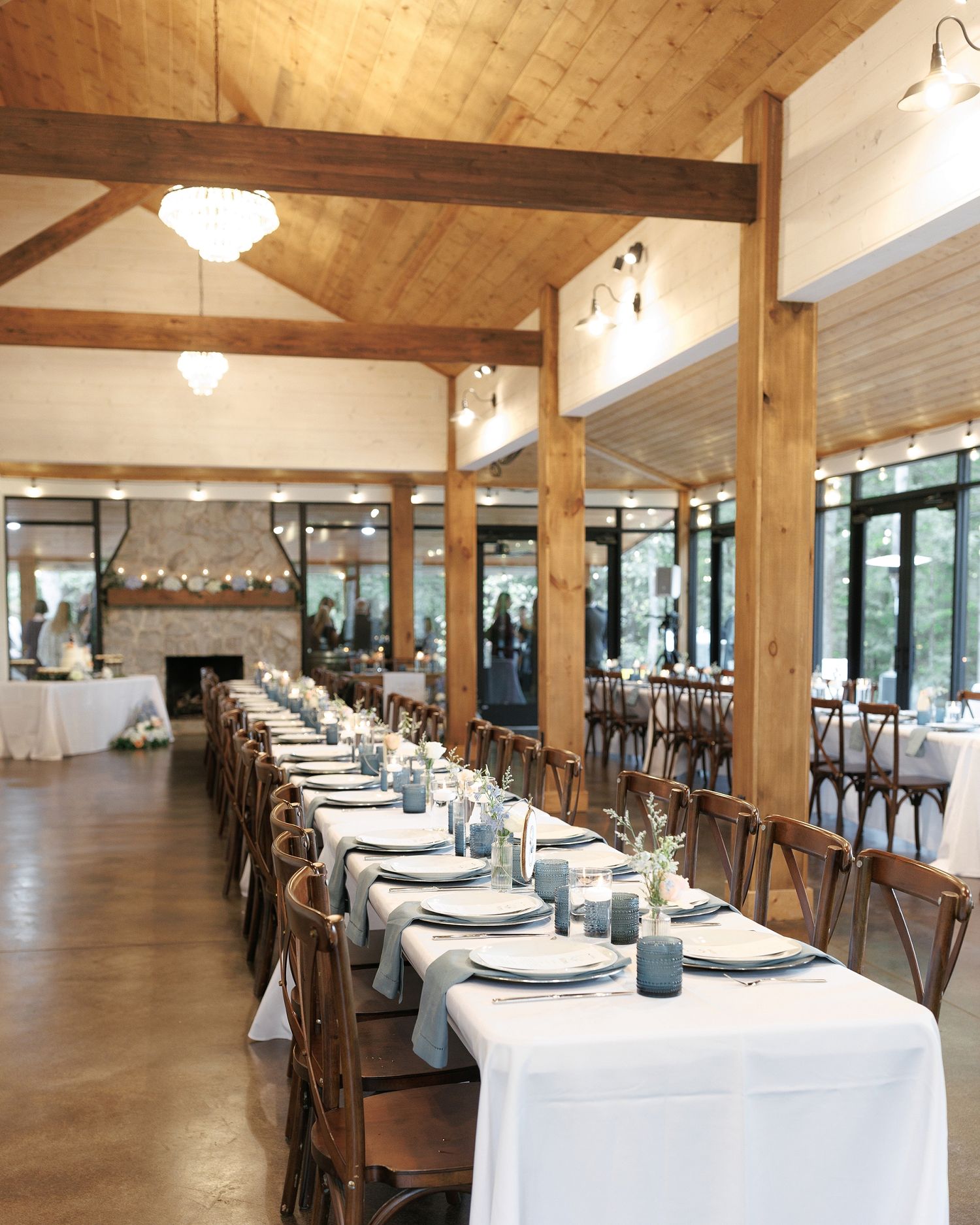 Elegant rustic wedding reception hall with long white tables, wooden beams, cross-back chairs, and blue glassware centerpieces.