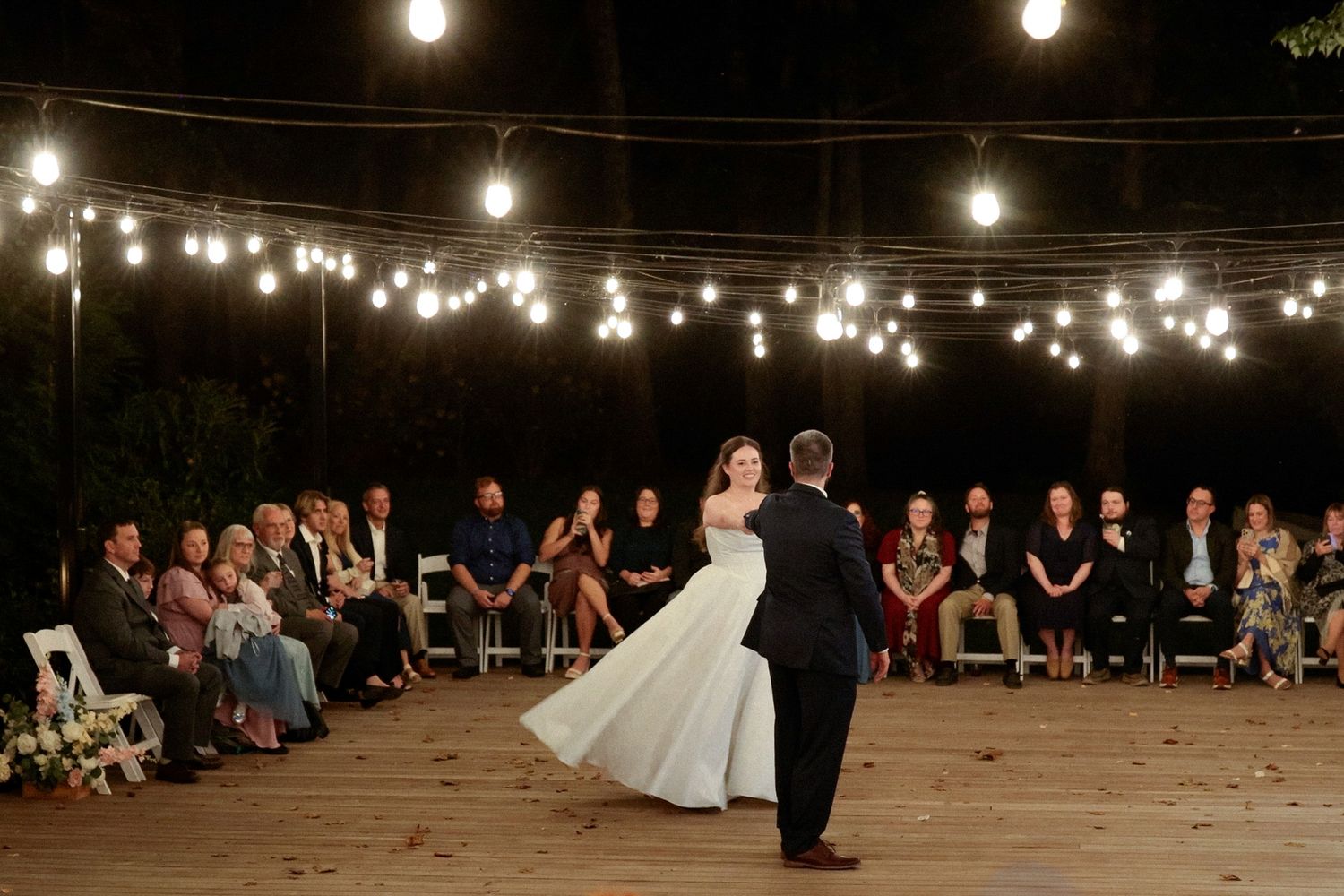 Bride and groom share first dance on outdoor wooden floor under string lights with seated wedding guests watching at night.