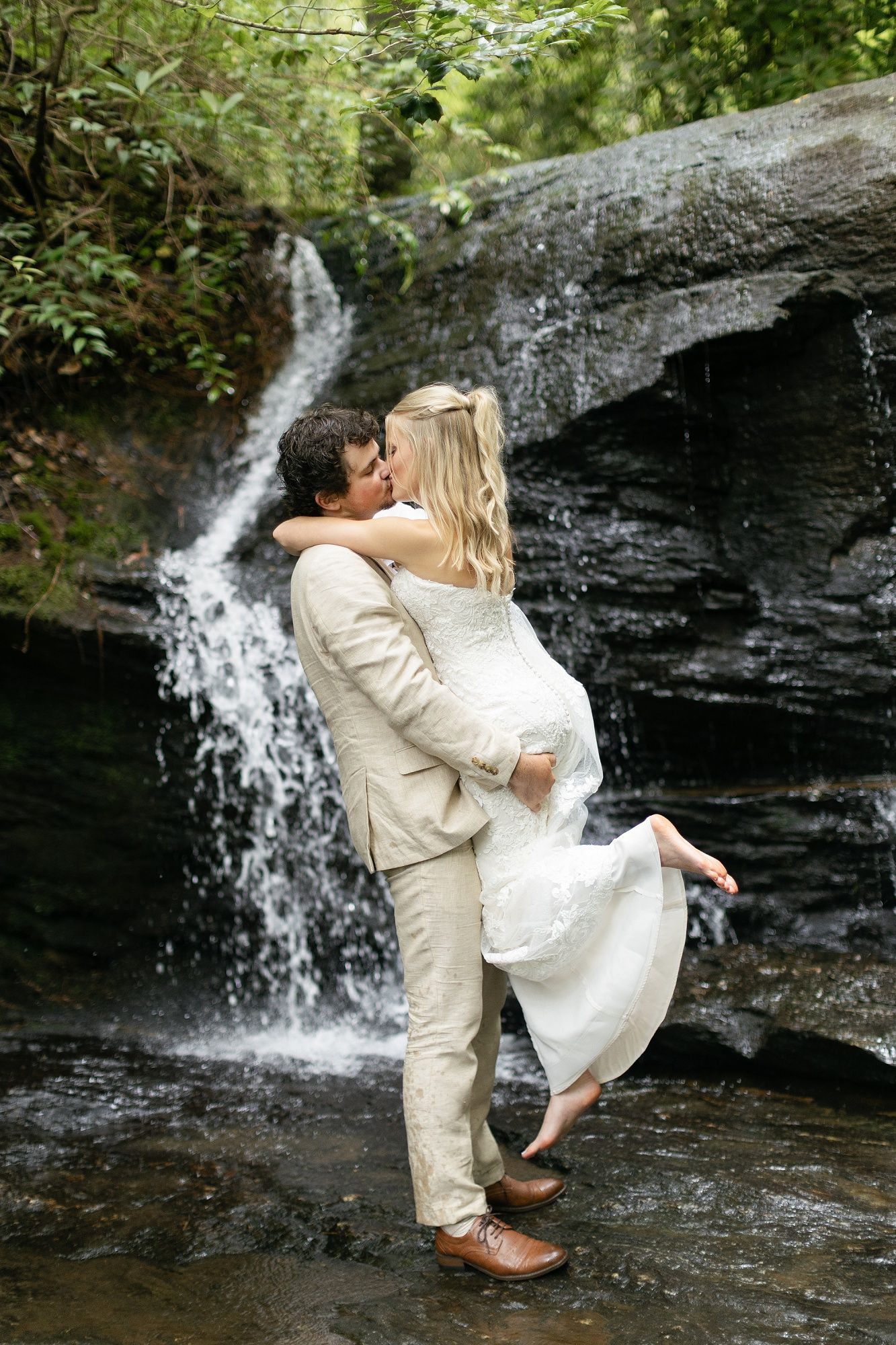 A romantic couple embraces by a cascading waterfall, the bride in white and groom in beige share a tender moment.
