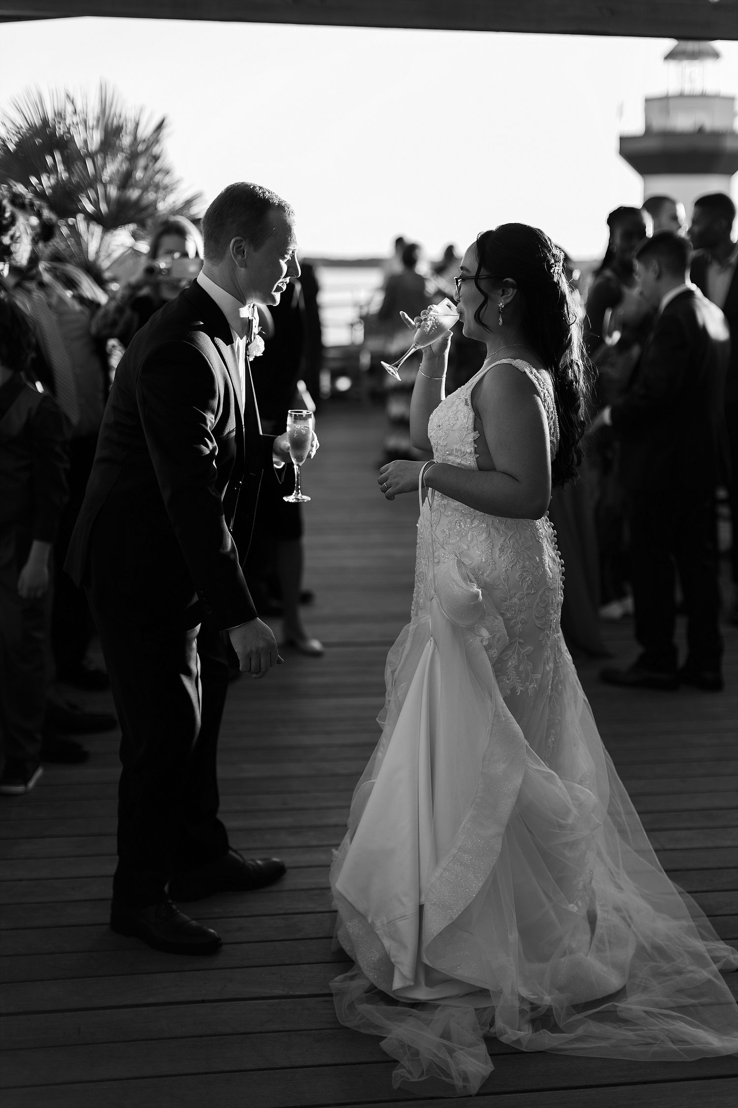 A black and white photo of a couple toasting with champagne glasses on a wooden deck at sunset.