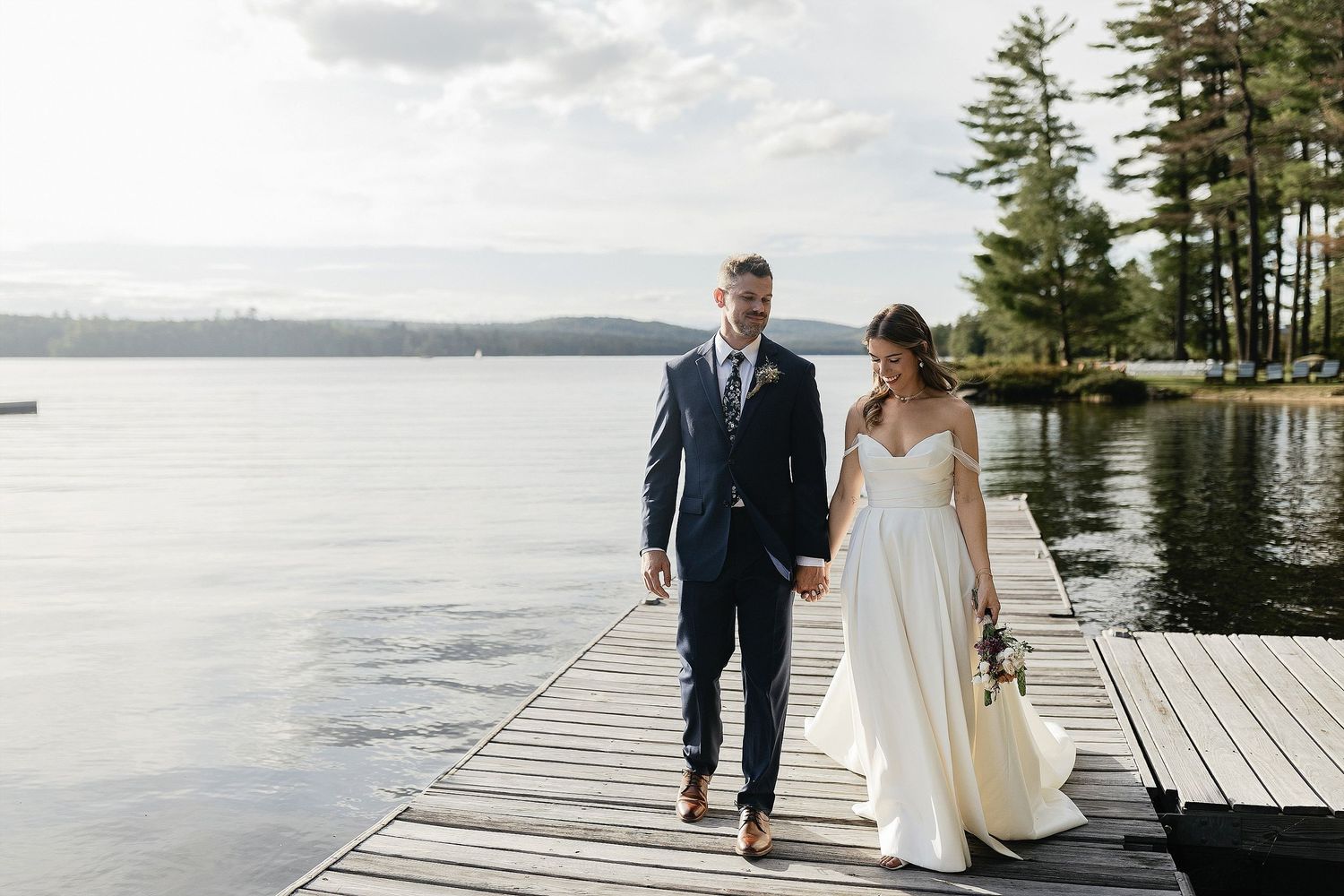 A couple walks together on a lakeside dock surrounded by pine trees and calm waters.