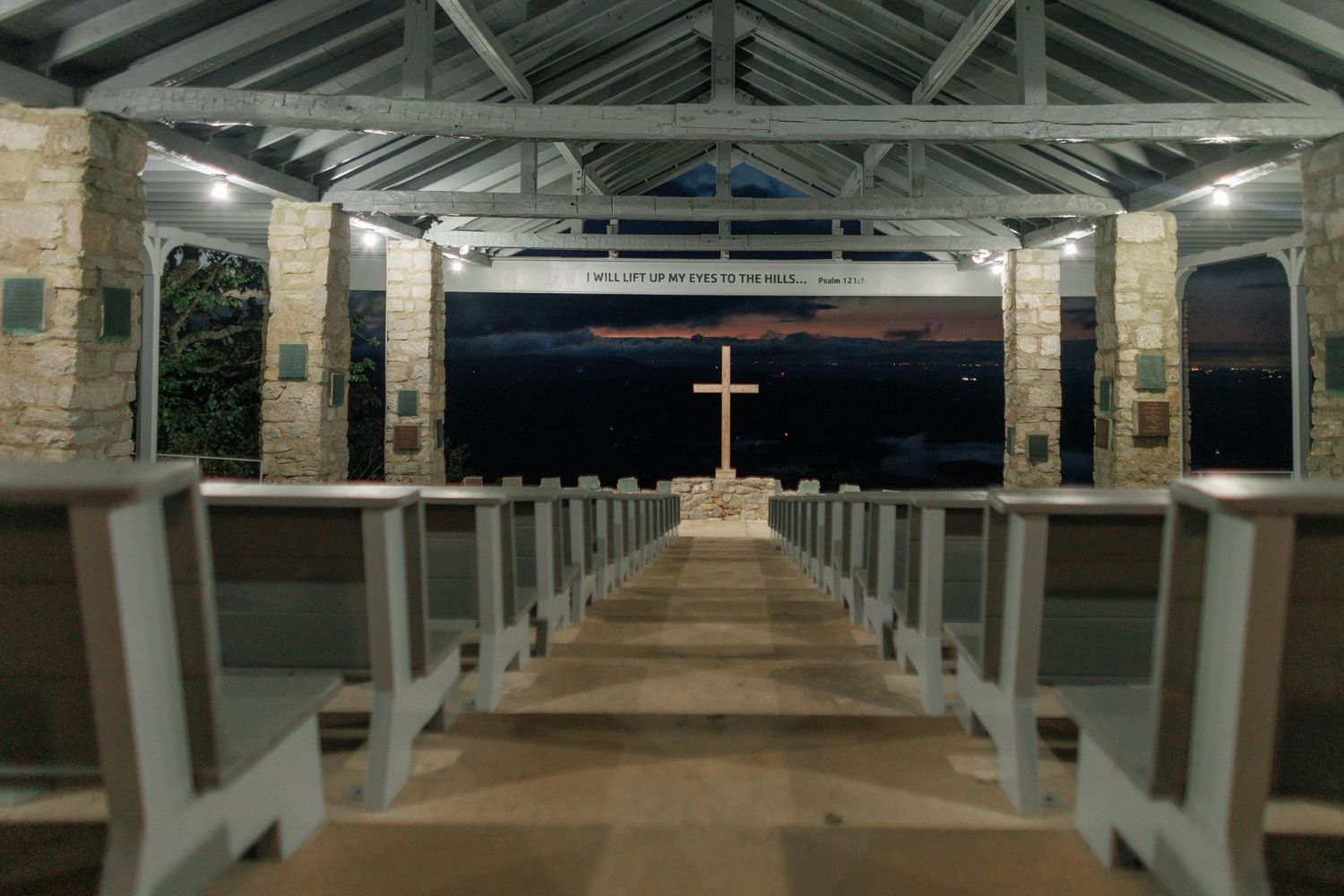An illuminated cross stands at the end of a long aisle lined with wooden pews inside a rustic stone church at night.