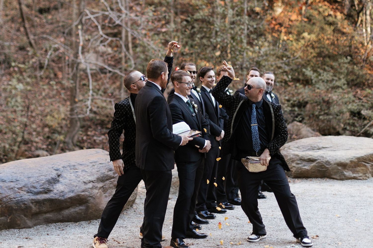 A group in formal black suits stands in a line on a stone path surrounded by autumn trees.