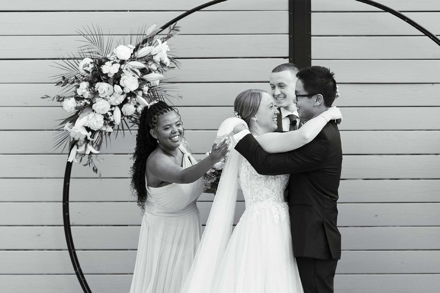Black and white photo of a wedding celebration with three people embracing in front of a circular floral arch backdrop.