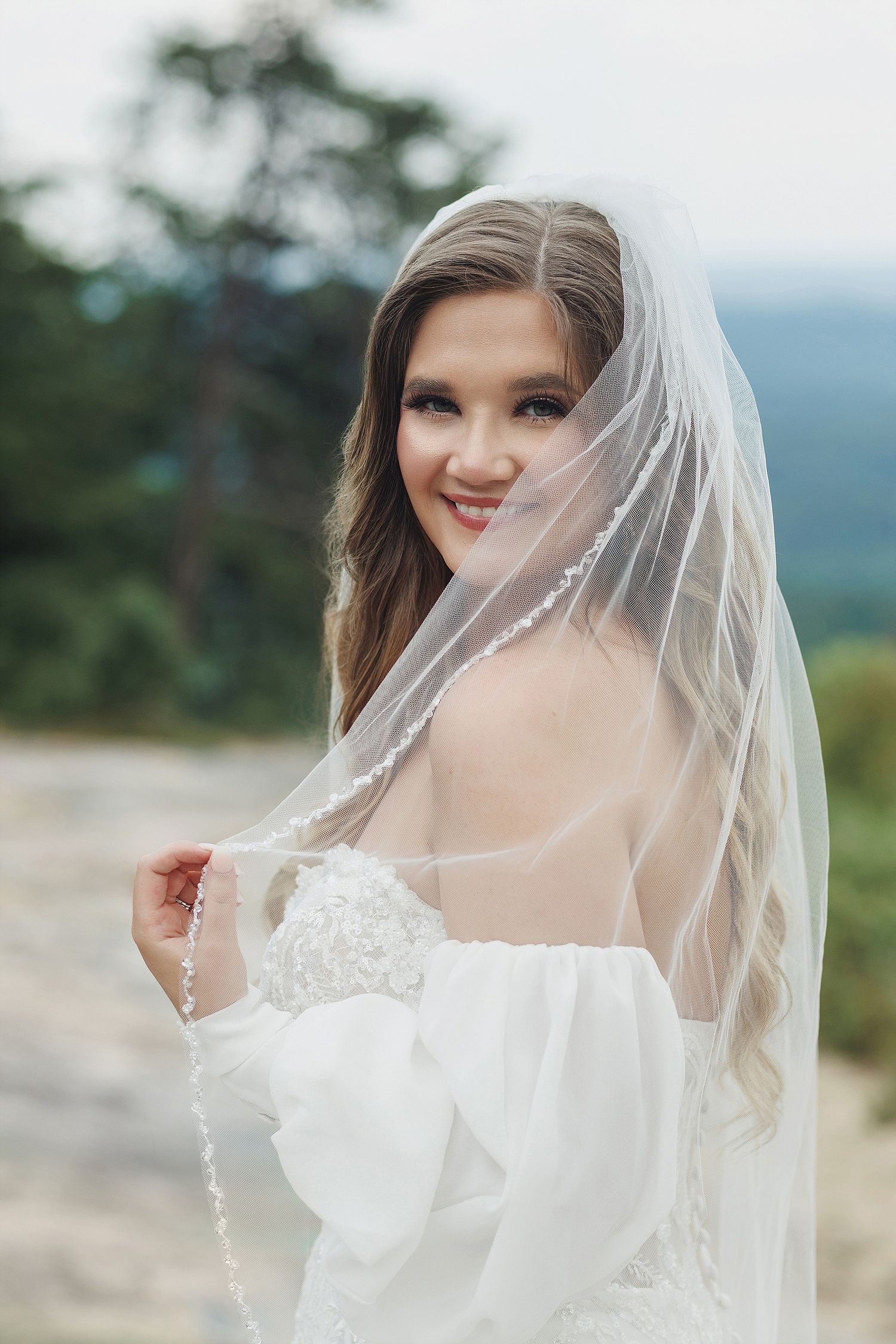 A bride wearing an off-shoulder wedding dress and veil poses outdoors with mountain scenery.