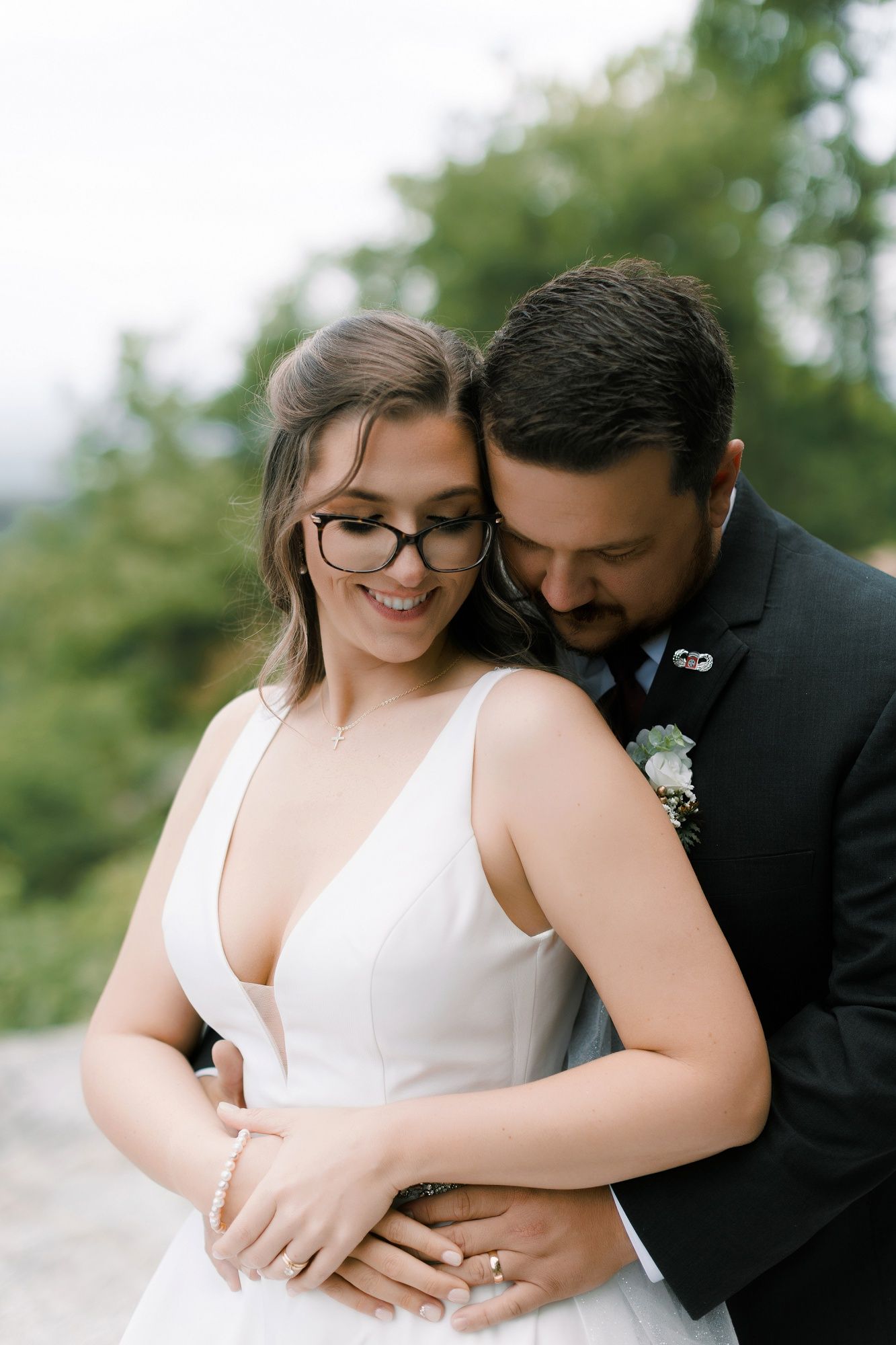 Groom embracing smiling bride wearing glasses in a white V-neck gown at an outdoor wedding.