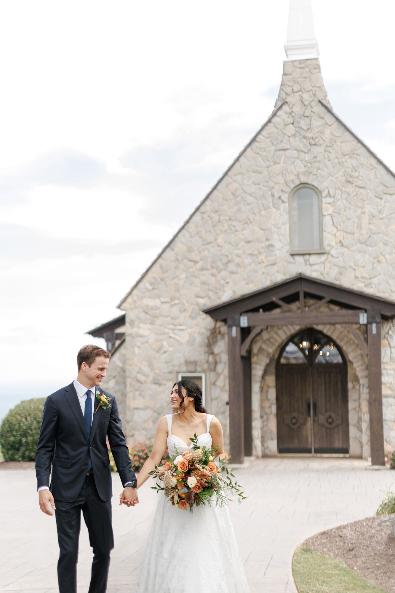 Bride and groom holding hands walking in front of a stone chapel on their wedding day.