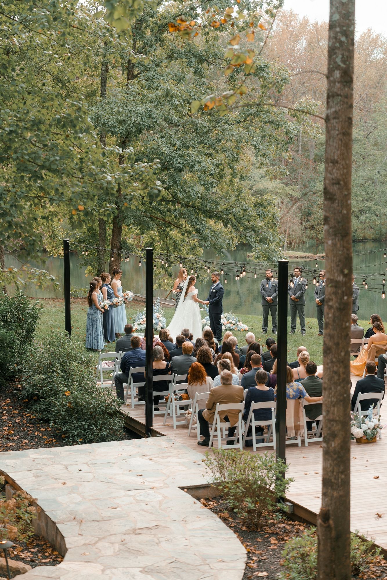 Outdoor wedding ceremony in a wooded setting with guests seated as bride and groom exchange vows near a pond.