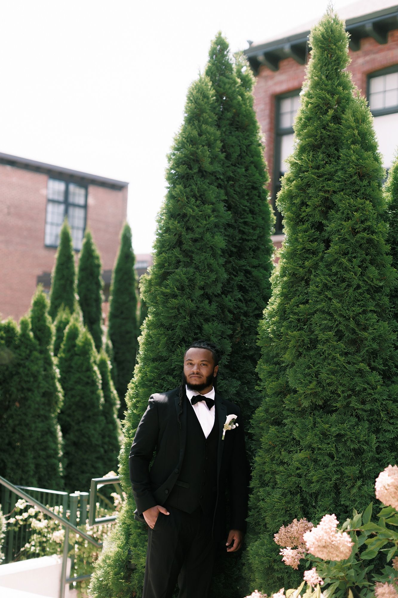 Groom in black tuxedo with bow tie and boutonniere stands among tall evergreen trees at outdoor wedding venue.