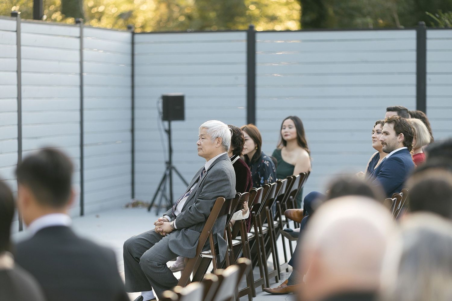 Wedding guests seated in rows at an outdoor ceremony with a white privacy fence backdrop and speaker stands.