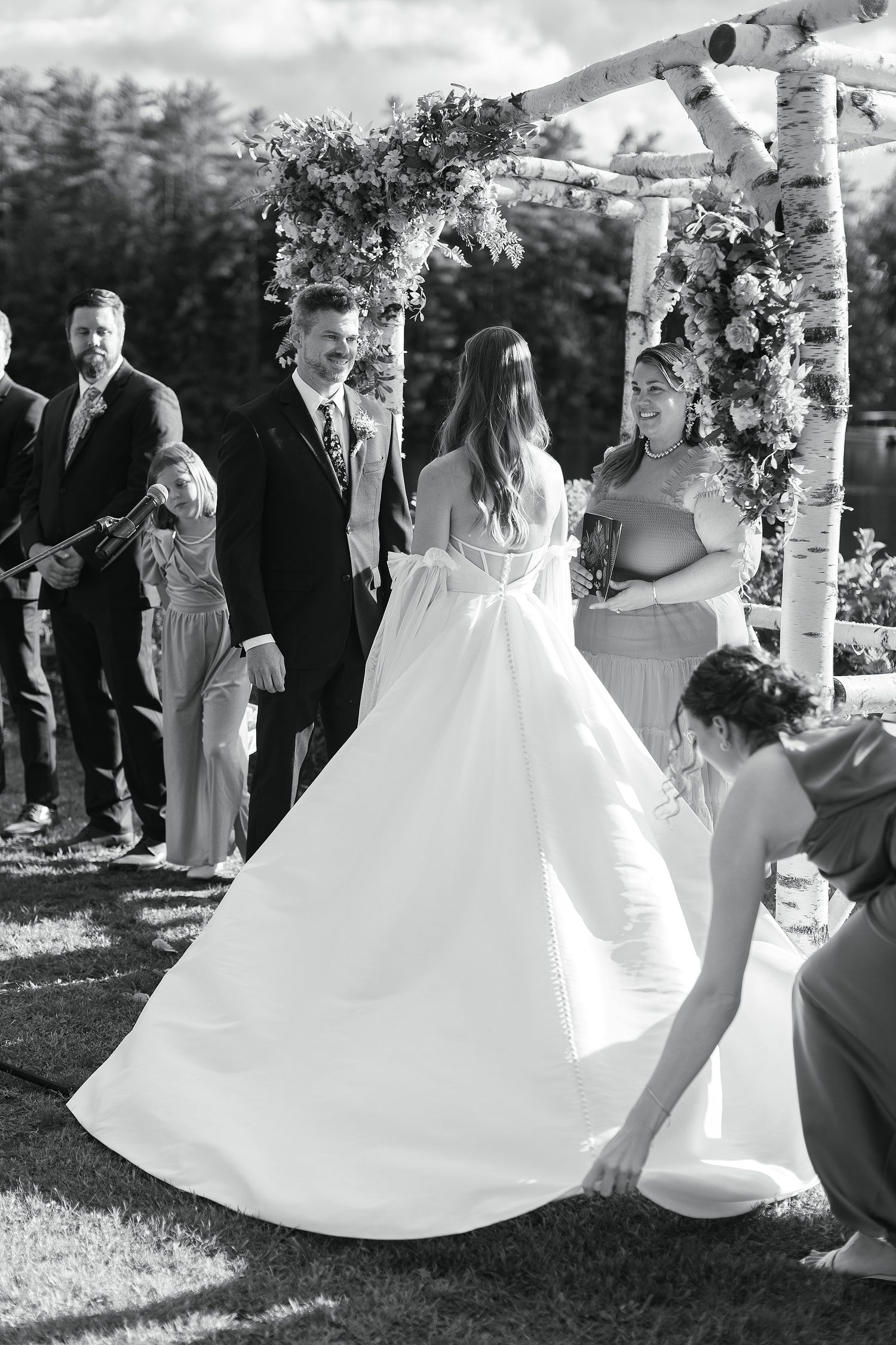 A wedding ceremony under a floral-decorated birch arch with guests seated on white chairs.