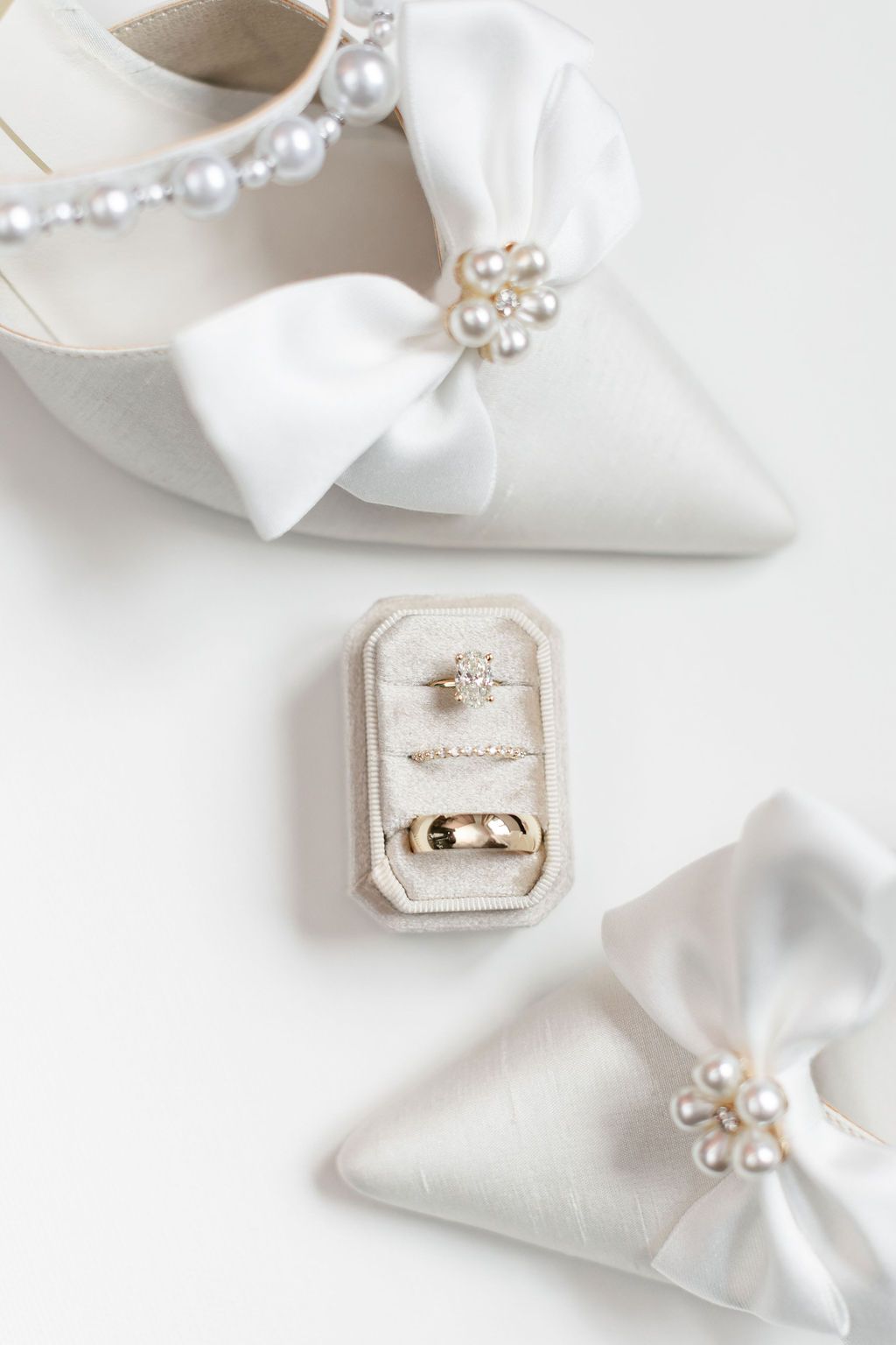 Elegant white bridal shoes with pearl details and satin bows displayed alongside wedding rings in a velvet ring box.