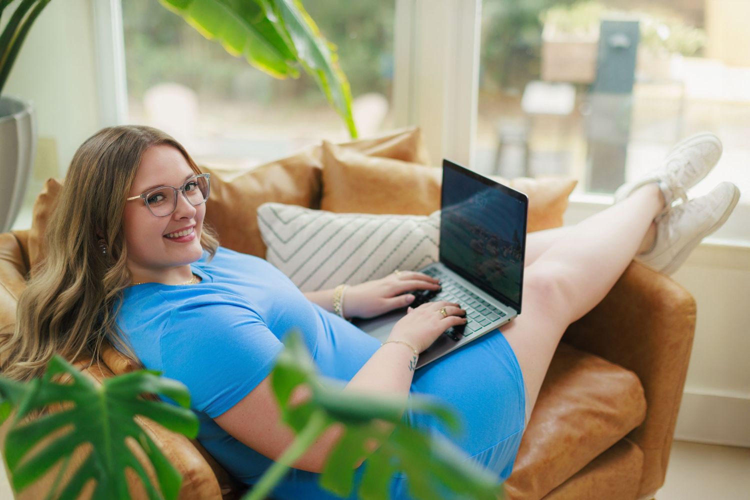 Woman in blue shirt smiling while using laptop on brown leather couch near window with plants.