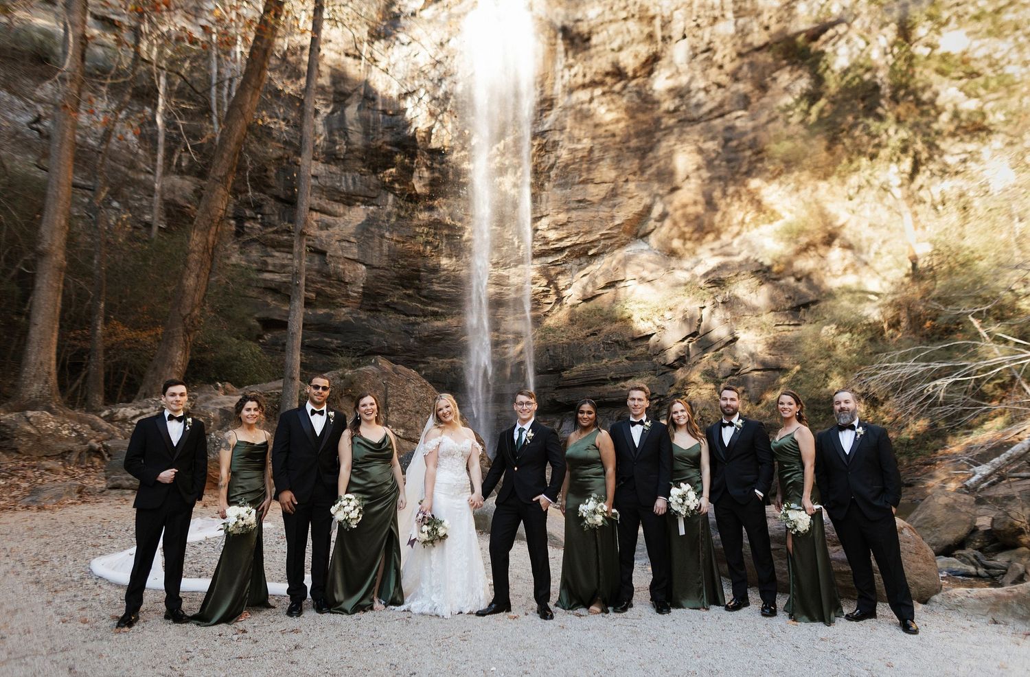 Wedding party poses in front of majestic natural waterfall with rock formations and trees in elegant formal attire.