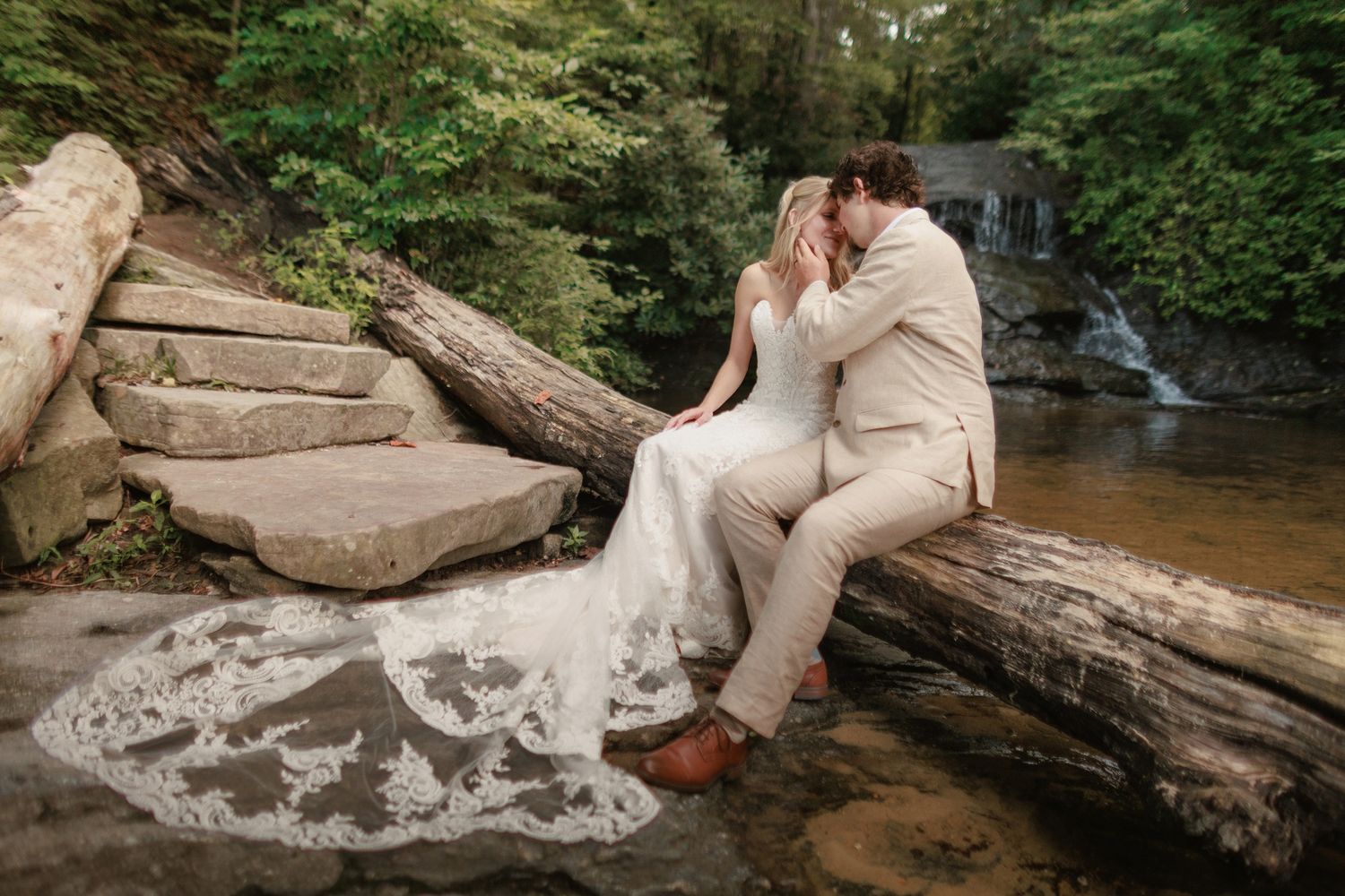 A couple in wedding attire shares a romantic moment on a fallen log near a forest waterfall.
