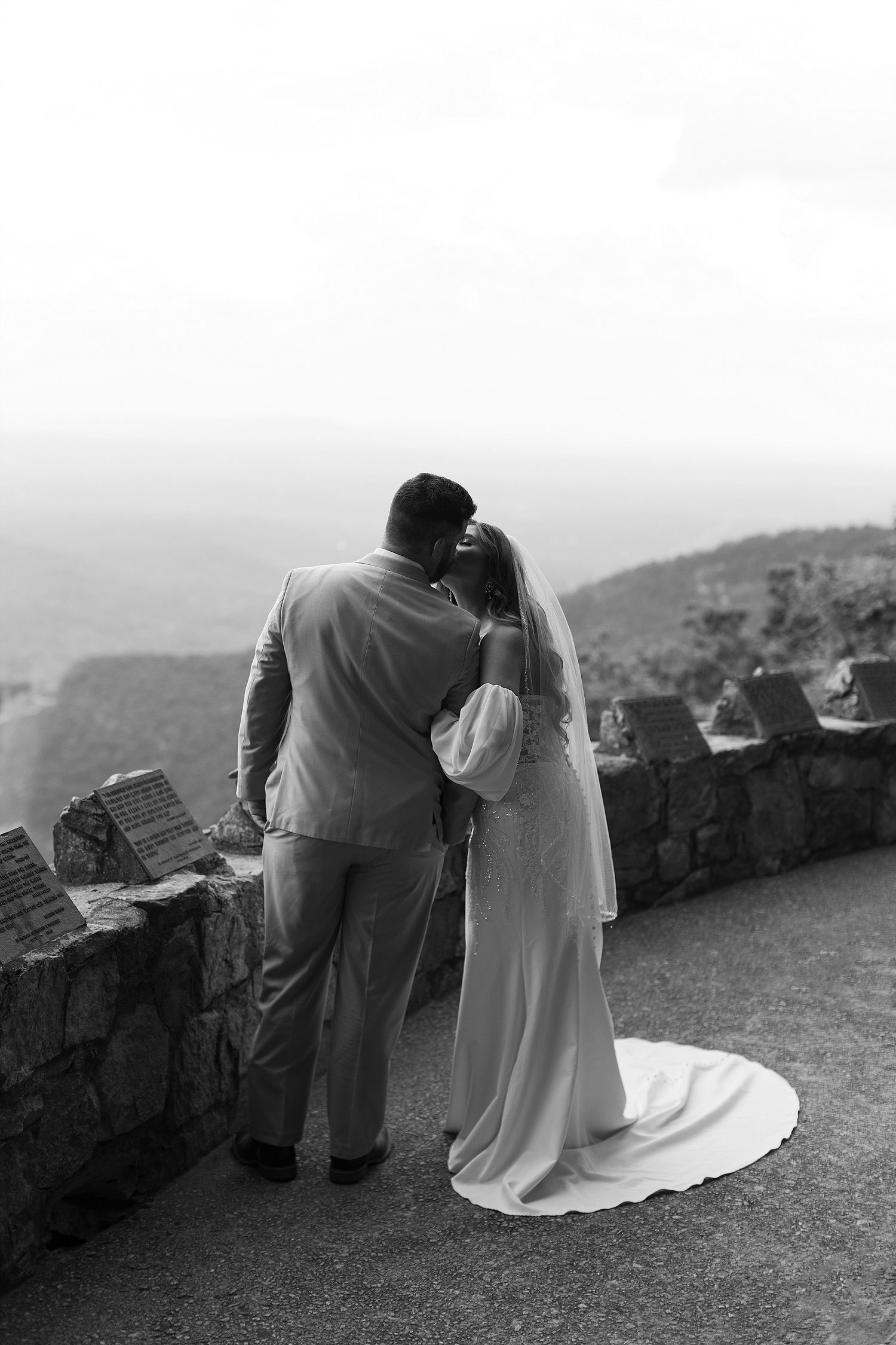Black and white photo of newlyweds looking out over mountain vista from stone overlook.