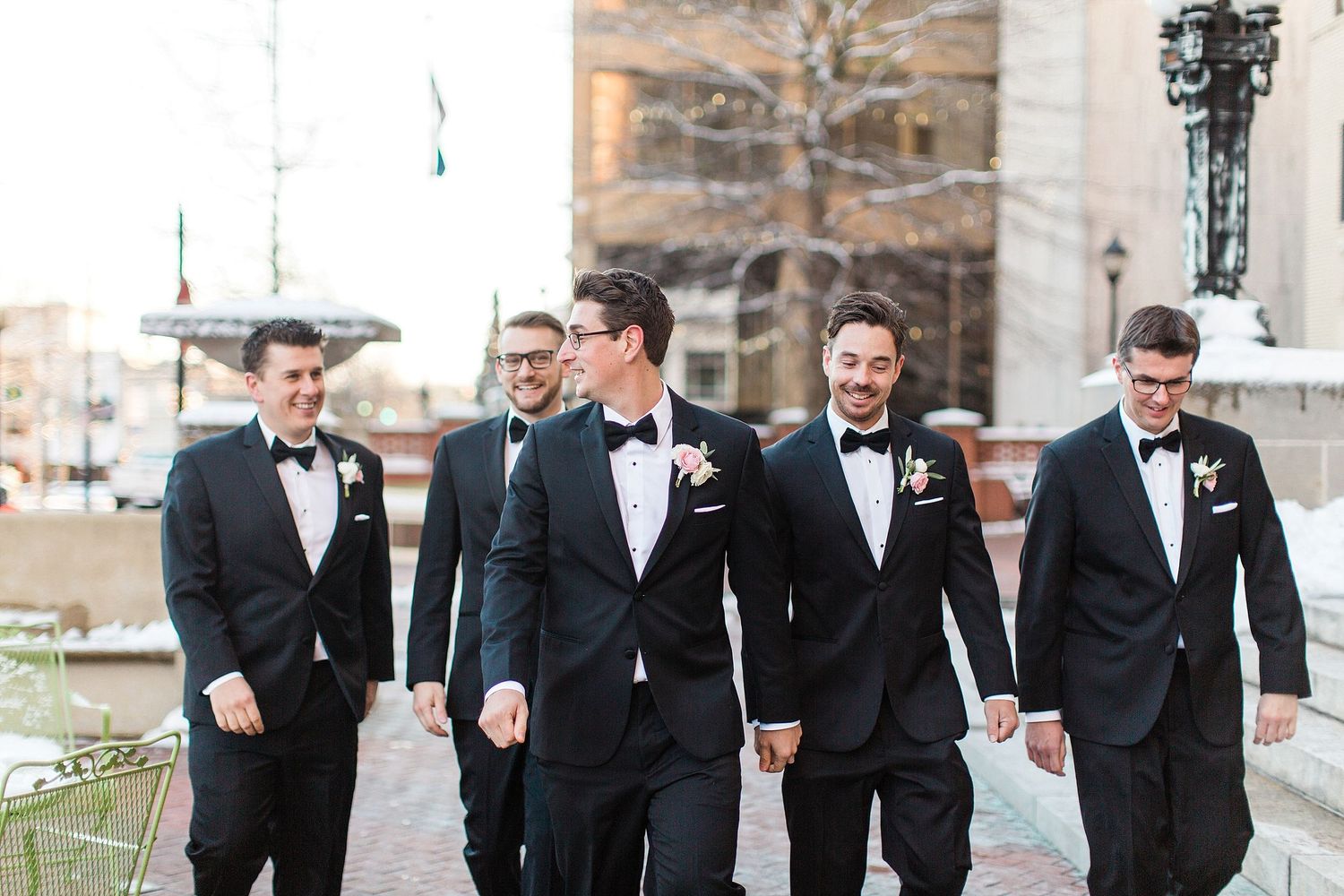 Groomsmen in black tuxedos walking and laughing together outdoors.