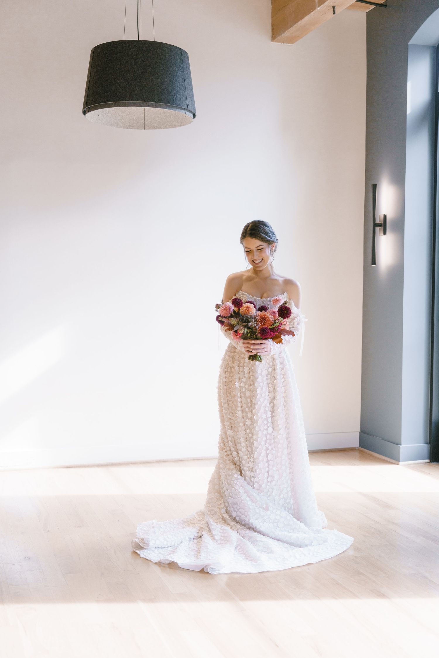 Bride in sparkly strapless wedding gown holding vibrant pink and red bouquet, standing in sunlit modern venue.