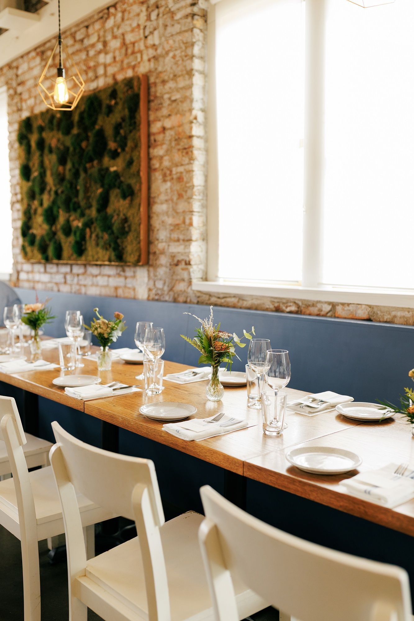Modern restaurant interior with exposed brick walls, white chairs and wooden tables set for dining service.