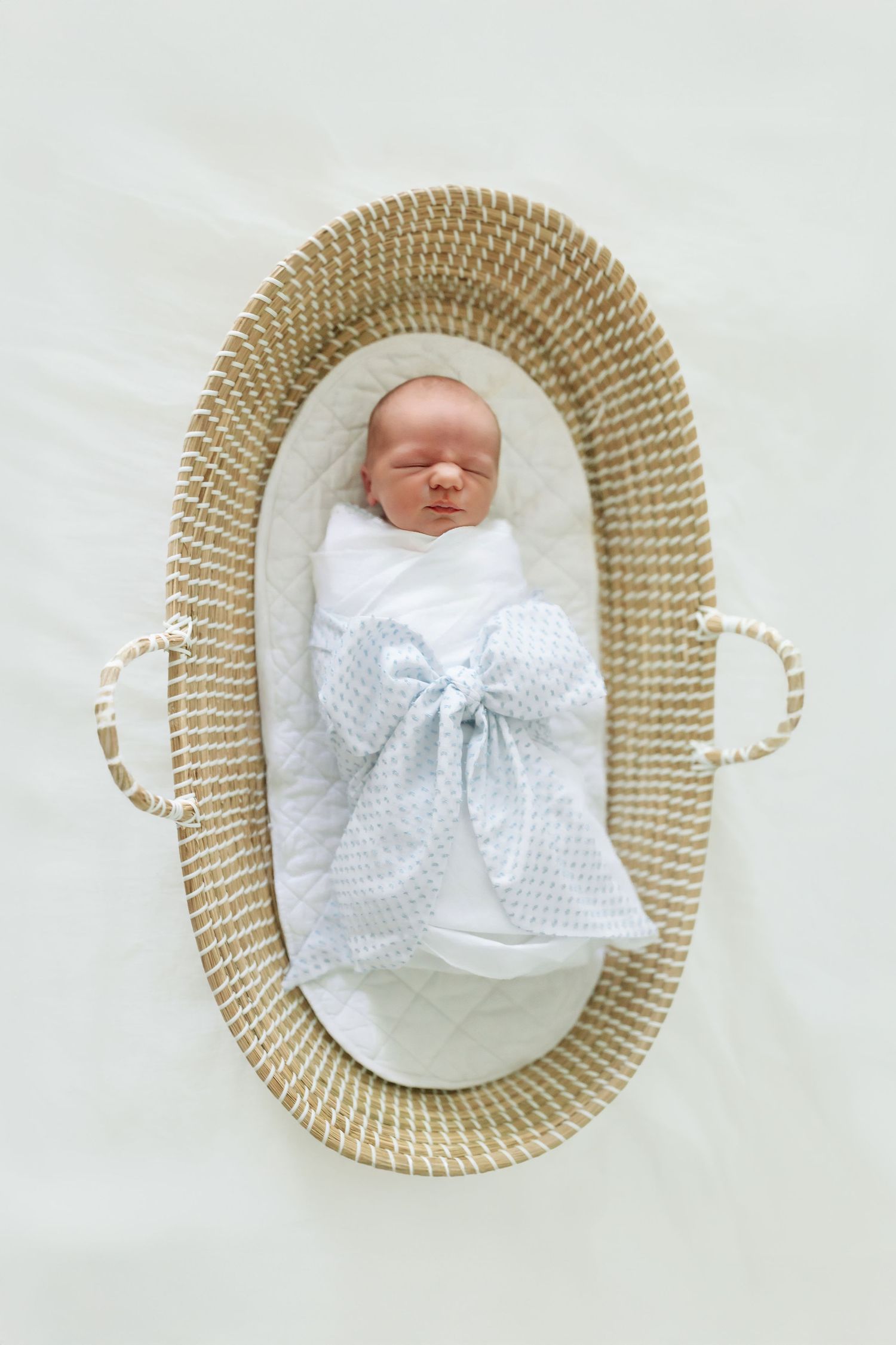 Newborn baby sleeping peacefully in a woven moses basket, swaddled in white with a light blue bow blanket.