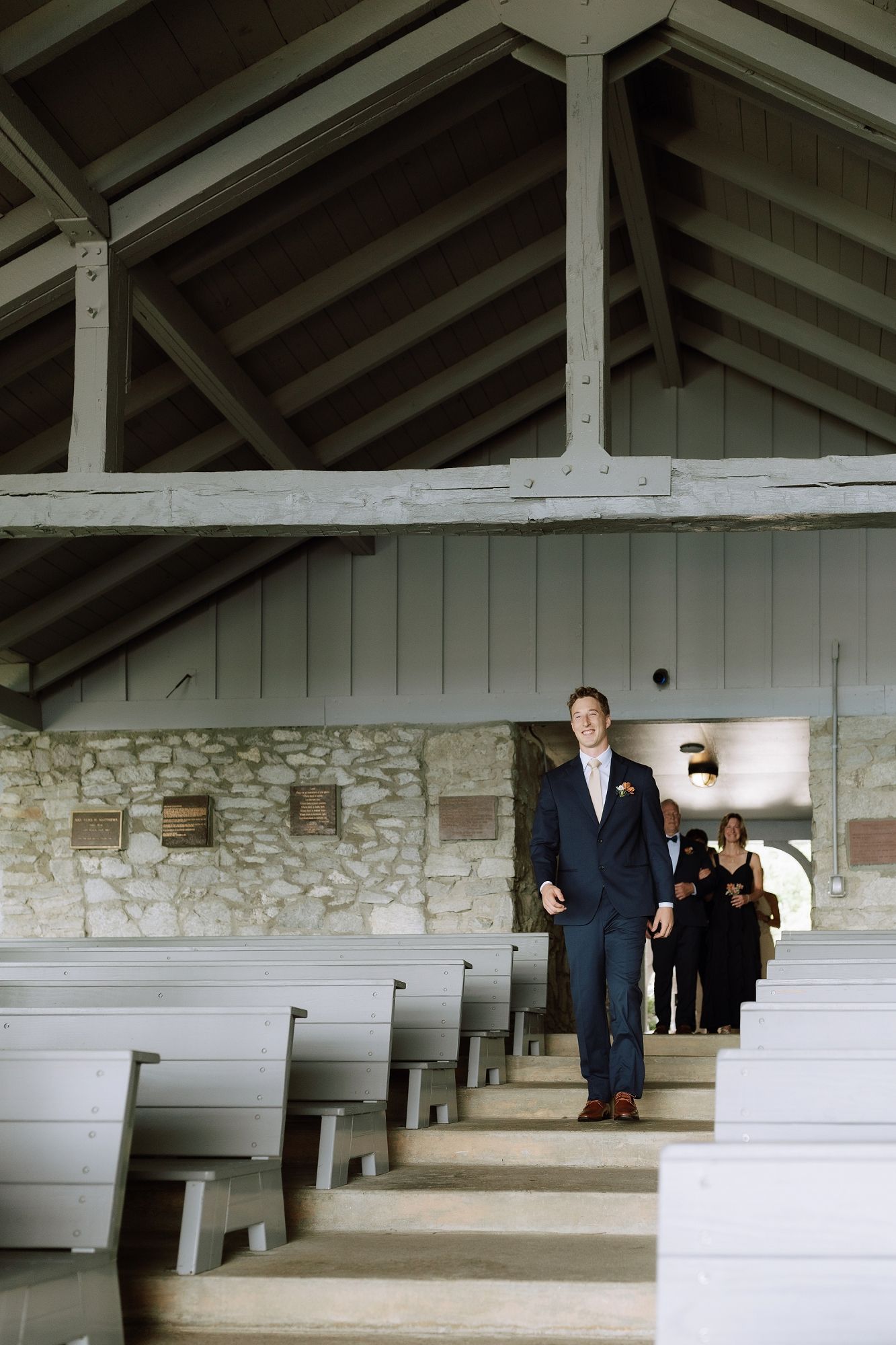 Groom walks down the aisle of a rustic stone chapel with wooden beam ceilings during a wedding ceremony.