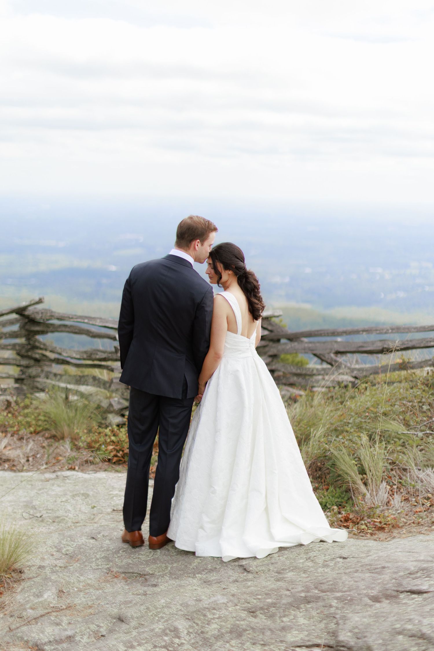 Bride and groom stand together on a mountain overlook, sharing an intimate moment in wedding attire.