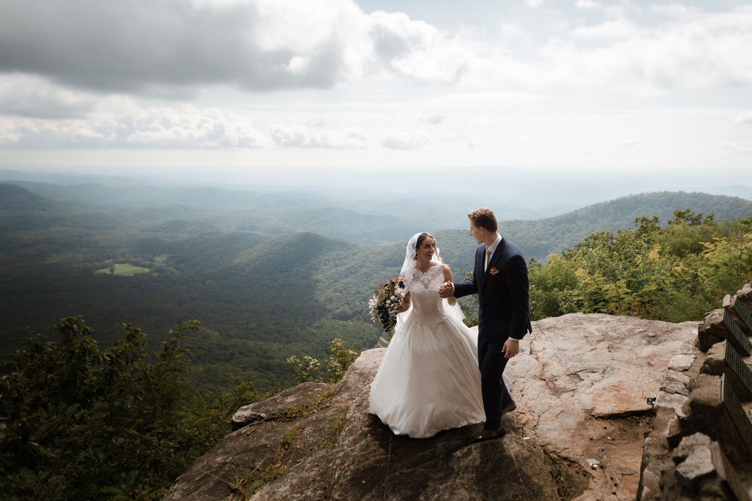 Bride and groom hold hands on a rocky mountain summit with sweeping green valley views below.