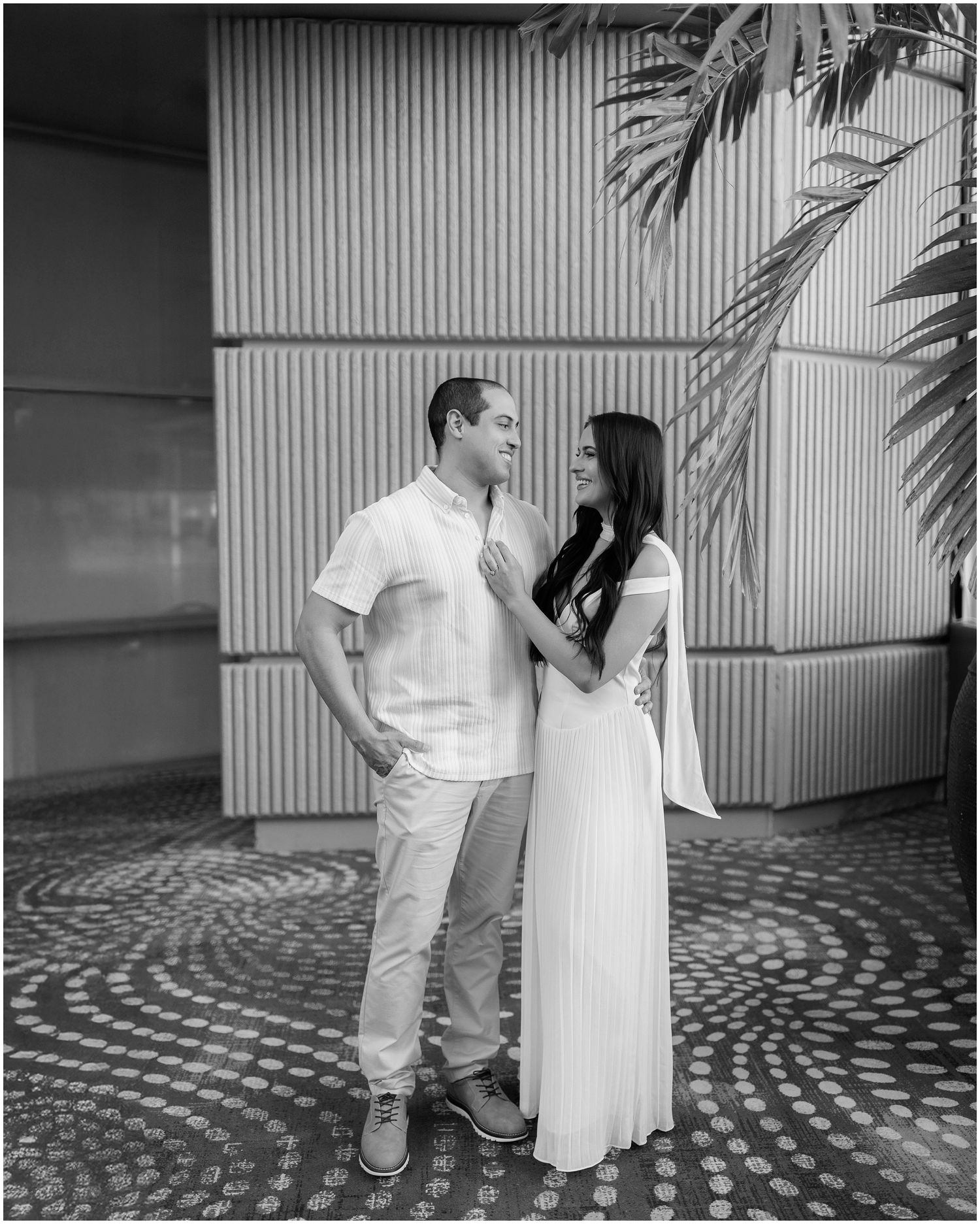 Black and white portrait of a couple in coordinated white outfits standing together by a textured wall.