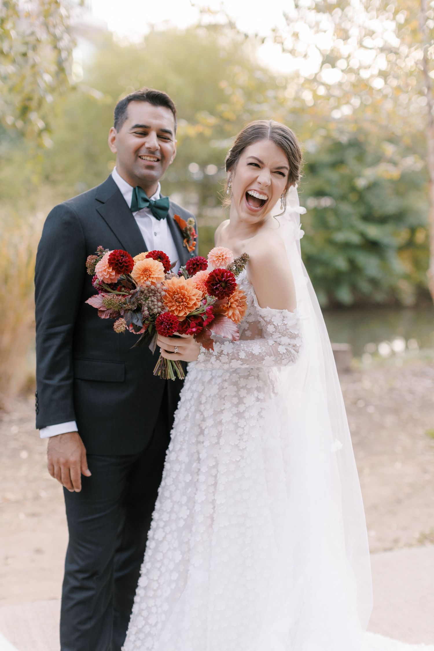 Joyful bride in white floral gown holding autumn bouquet stands with groom in black tuxedo outdoors.