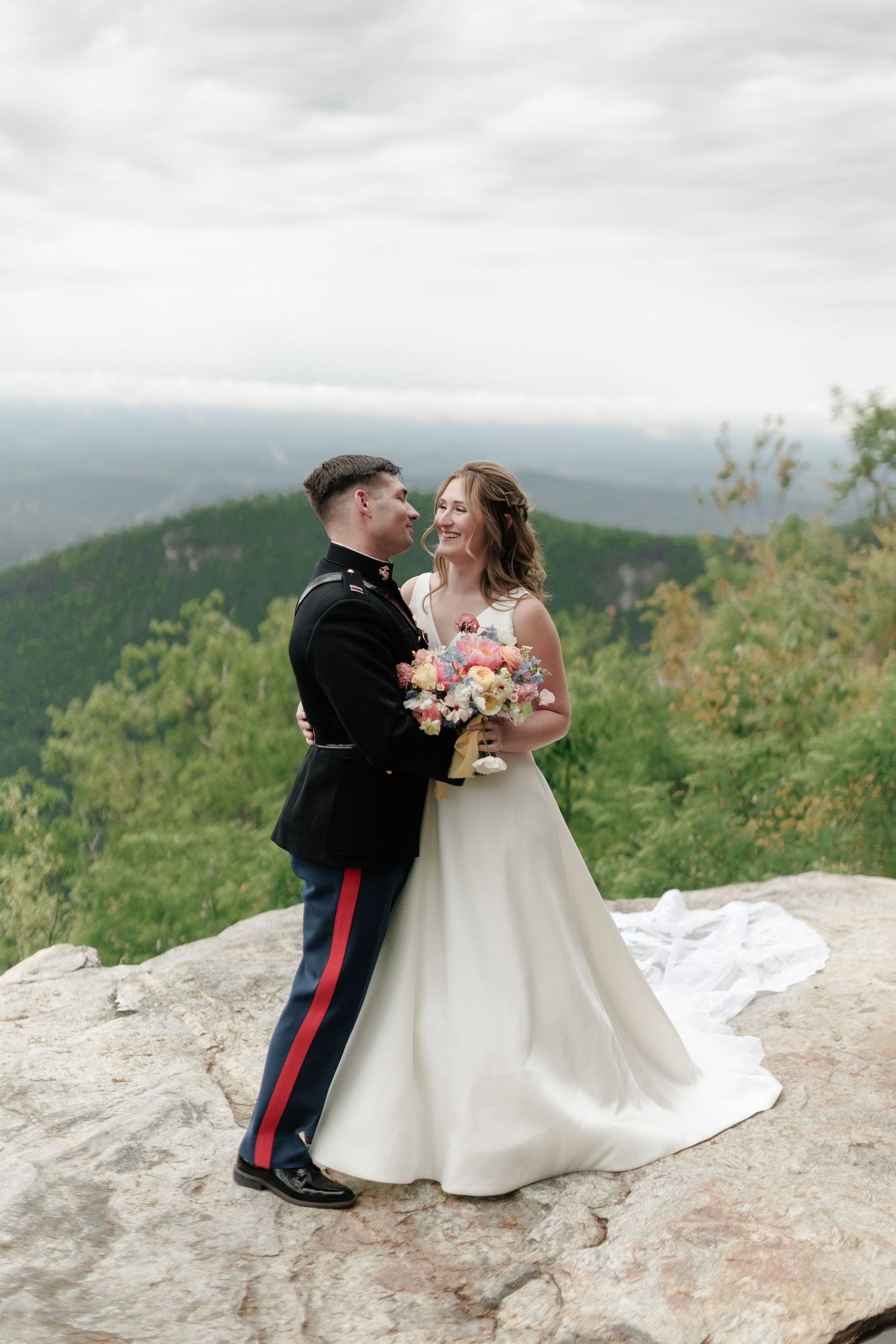 A wedding couple stands together on a mountaintop overlook with scenic forest views in the background on a cloudy day.