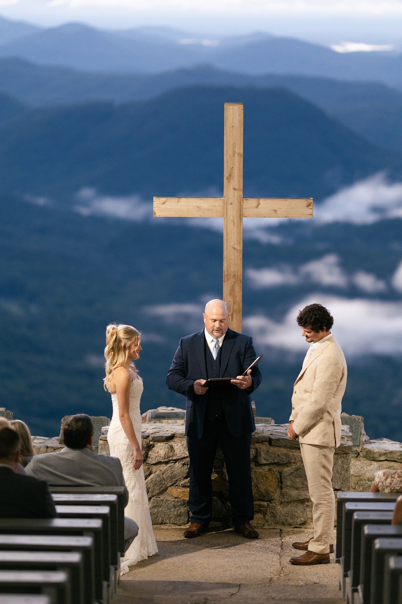 A mountain wedding ceremony takes place on a scenic overlook with a wooden cross and misty blue ridges in the background.