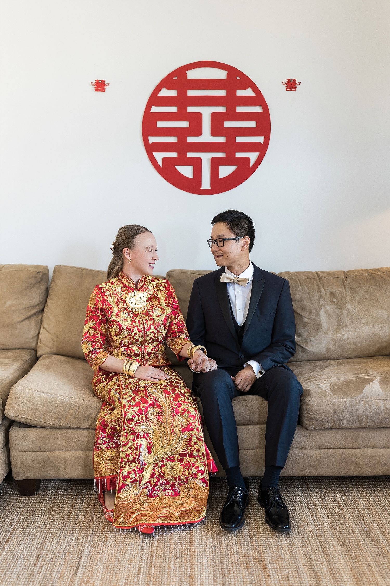 Couple sits on tan couch beneath red Chinese double happiness symbol, wearing traditional wedding attire.