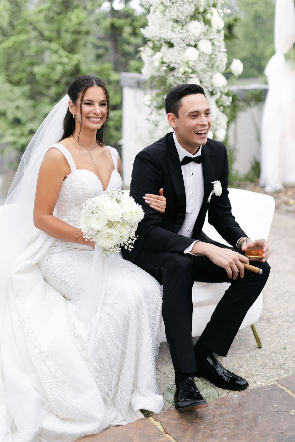 Newlyweds share a relaxed moment on white furniture with floral arch in background.