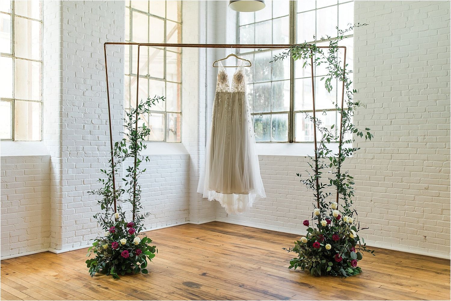 White wedding dress hanging from copper arch with floral arrangements in bright sunlit room.