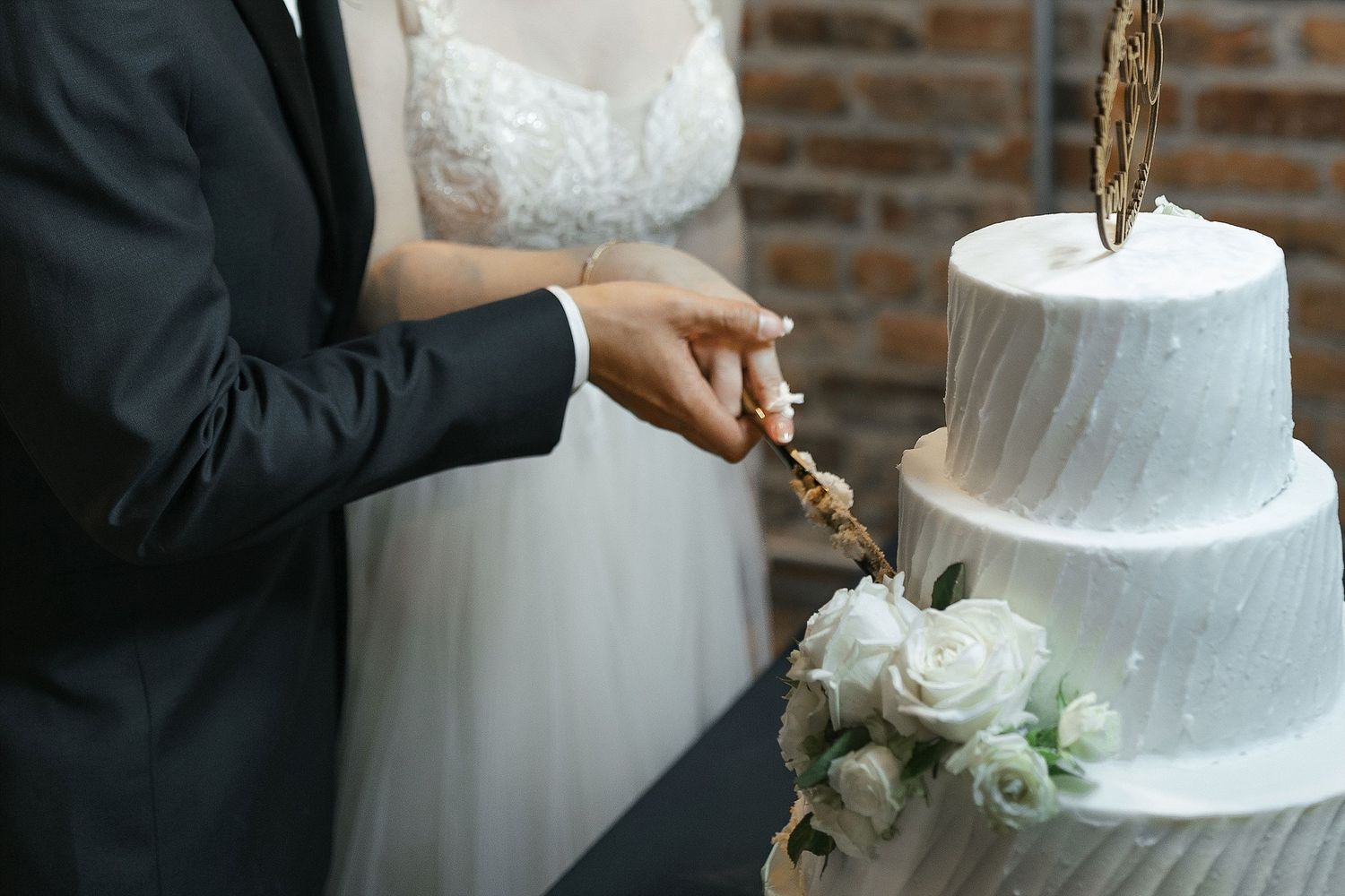 Two people cutting into an elegant white two-tier wedding cake decorated with fresh roses at a reception.