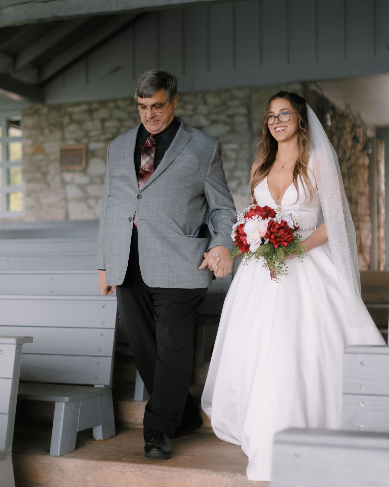 Father walks bride down the aisle in a rustic stone chapel as she carries a red and white bouquet.