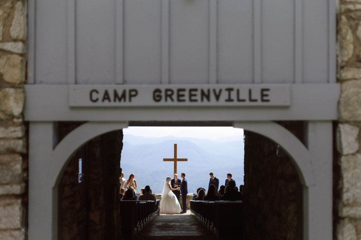 Wedding ceremony at Camp Greenville chapel with mountain views, bride and groom at altar with wooden cross backdrop.