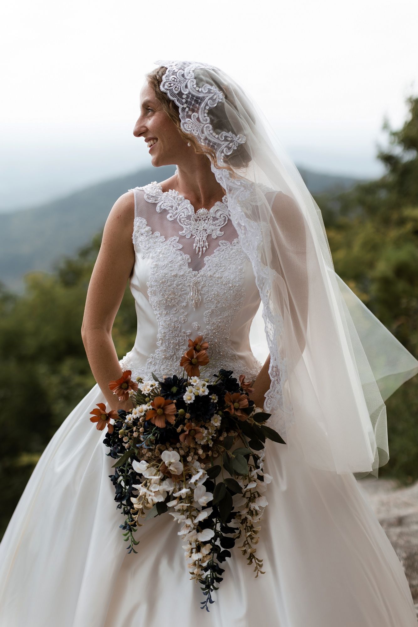 Smiling bride in lace wedding gown and veil holds cascading bouquet with mountain views behind her.