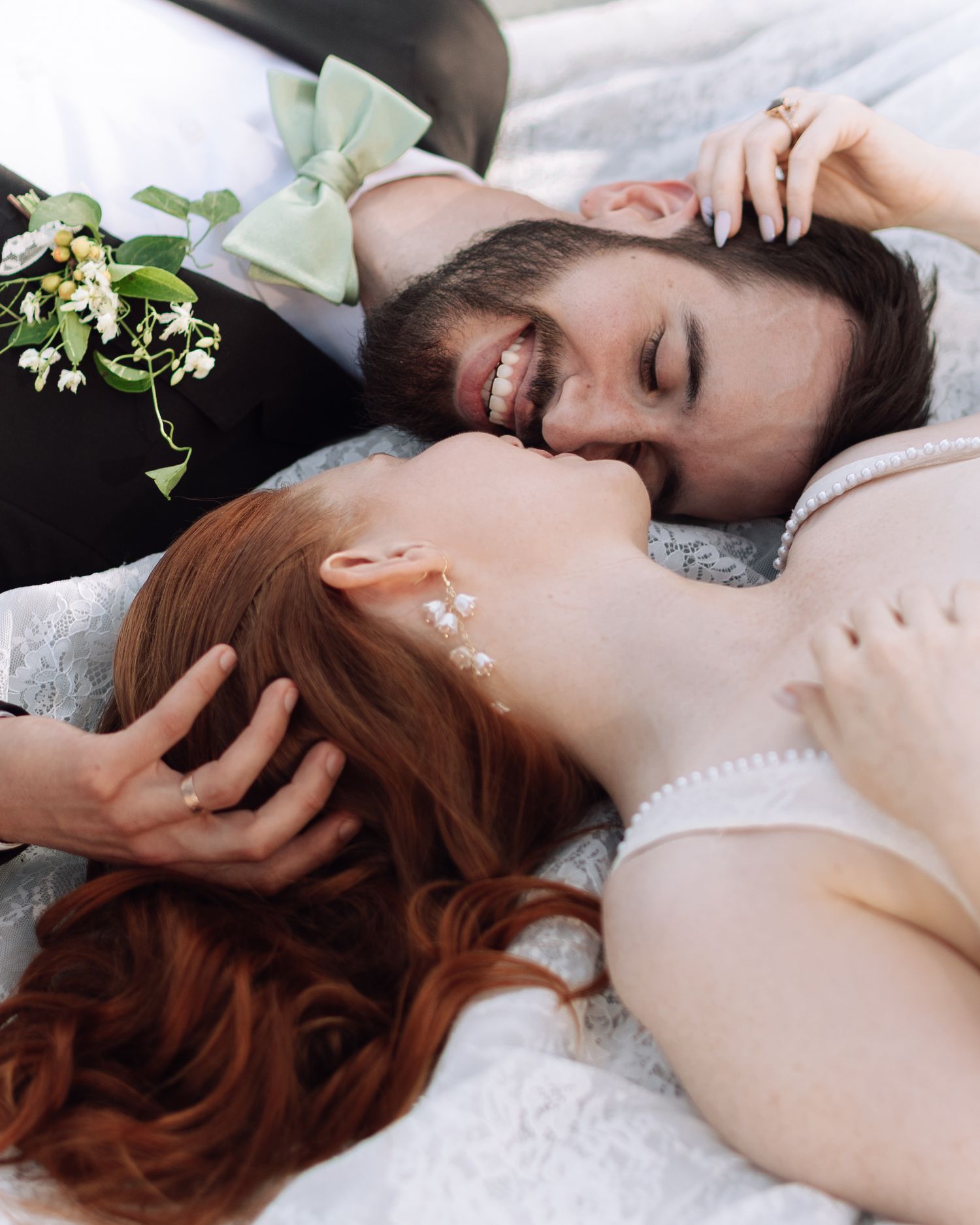 A smiling groom with a green bow tie and bride with red hair share a tender moment lying together on their wedding day.