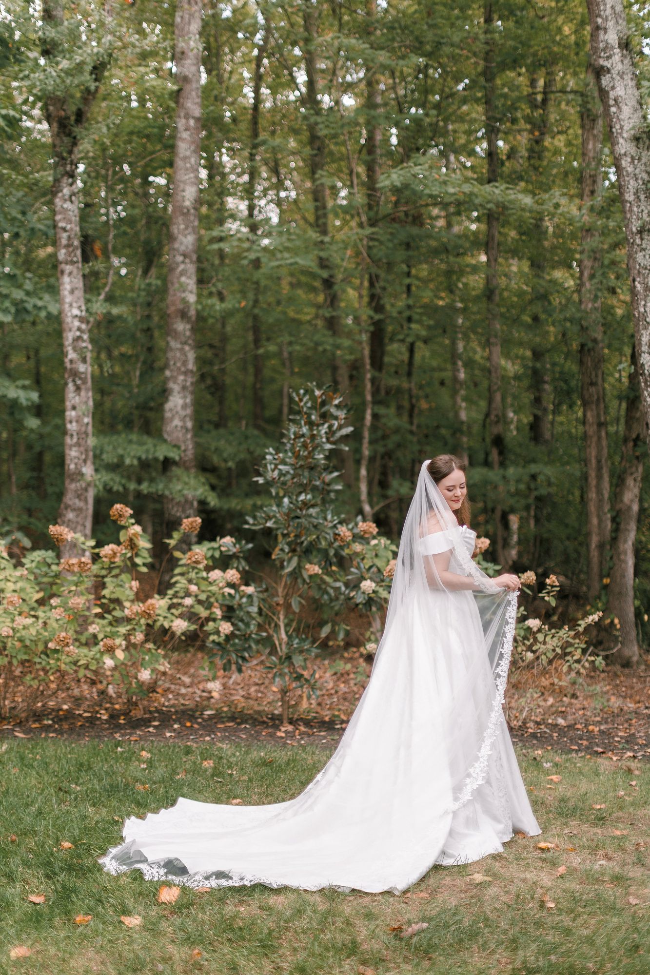 Elegant bride in white wedding gown with long lace veil standing in autumn garden with trees.