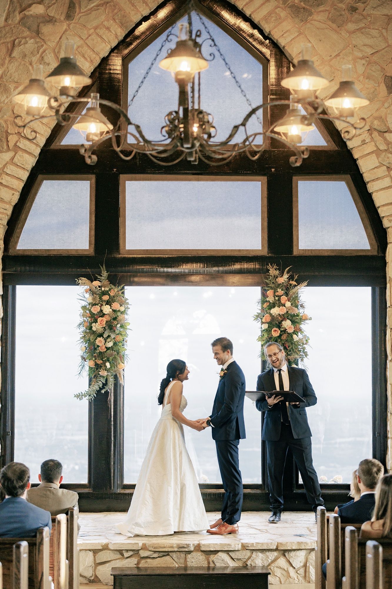 Wedding ceremony taking place in rustic chapel with stone walls, chandelier lighting and floral arch arrangements.