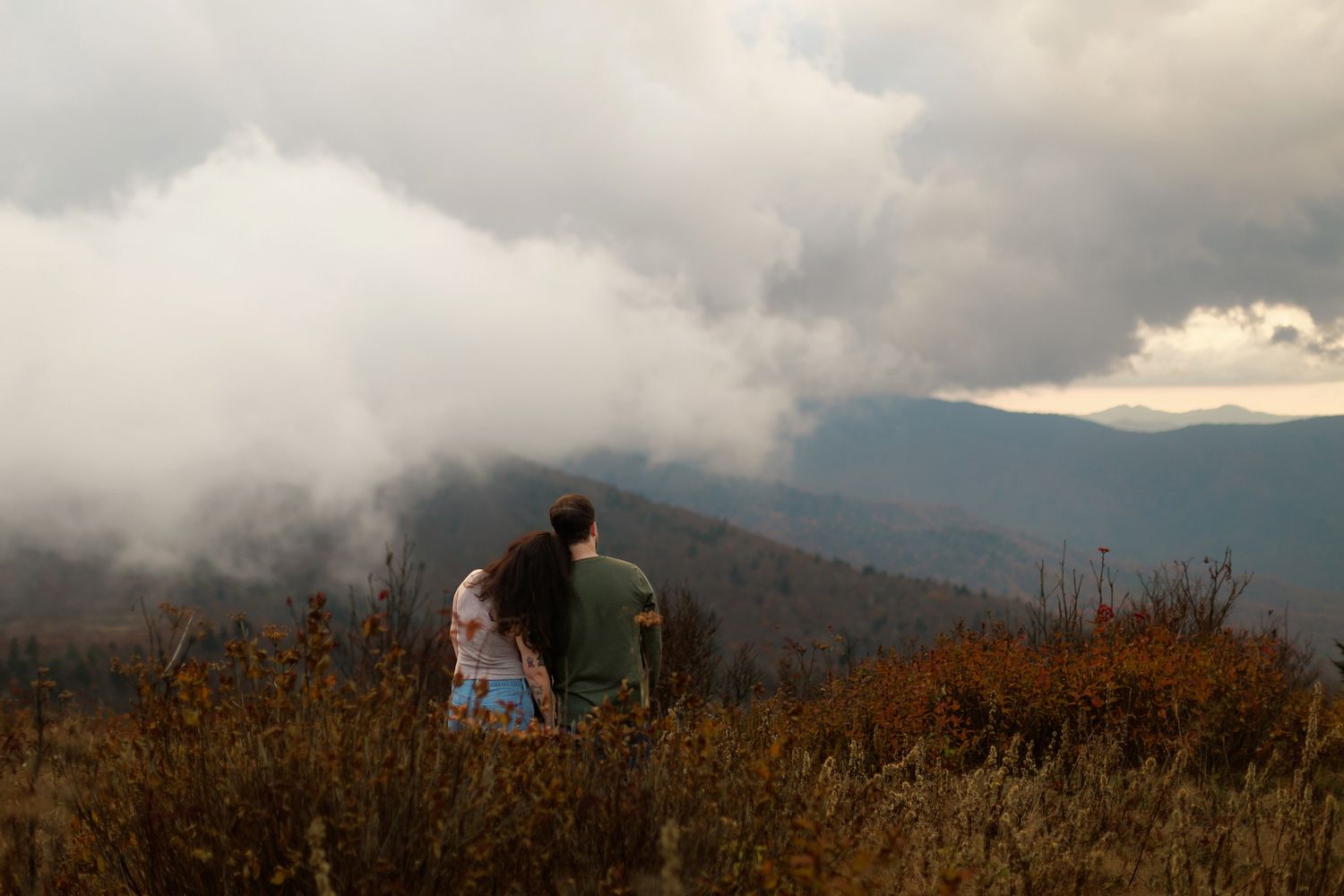 A couple sits together on a mountain overlook with dramatic cloudy skies and autumn landscape below.