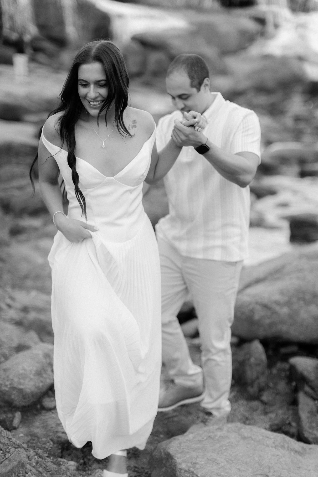 Black and white photo of a couple laughing together on rocky terrain wearing white outfits.