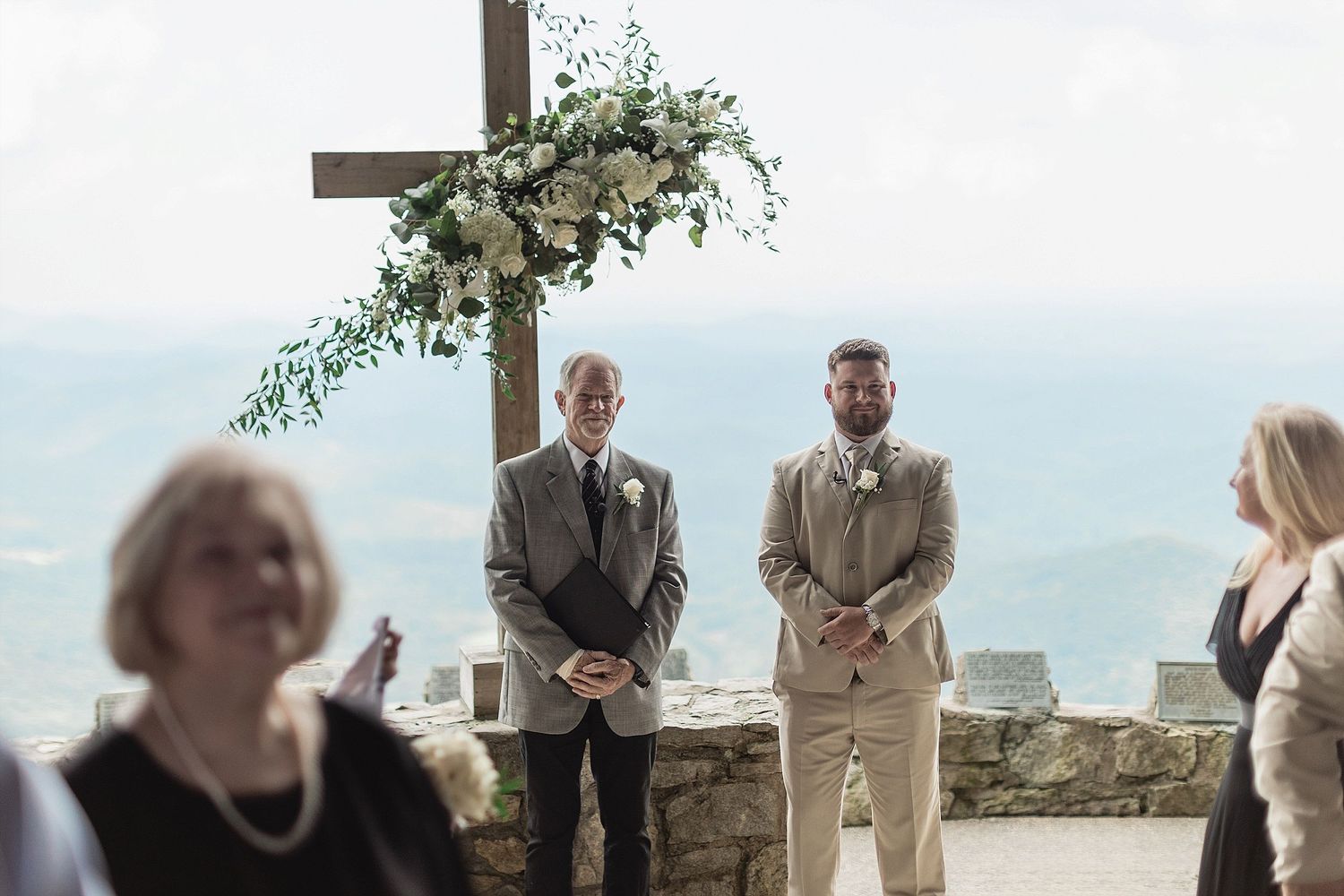 Outdoor wedding ceremony setup with floral cross arch and mountain vista.