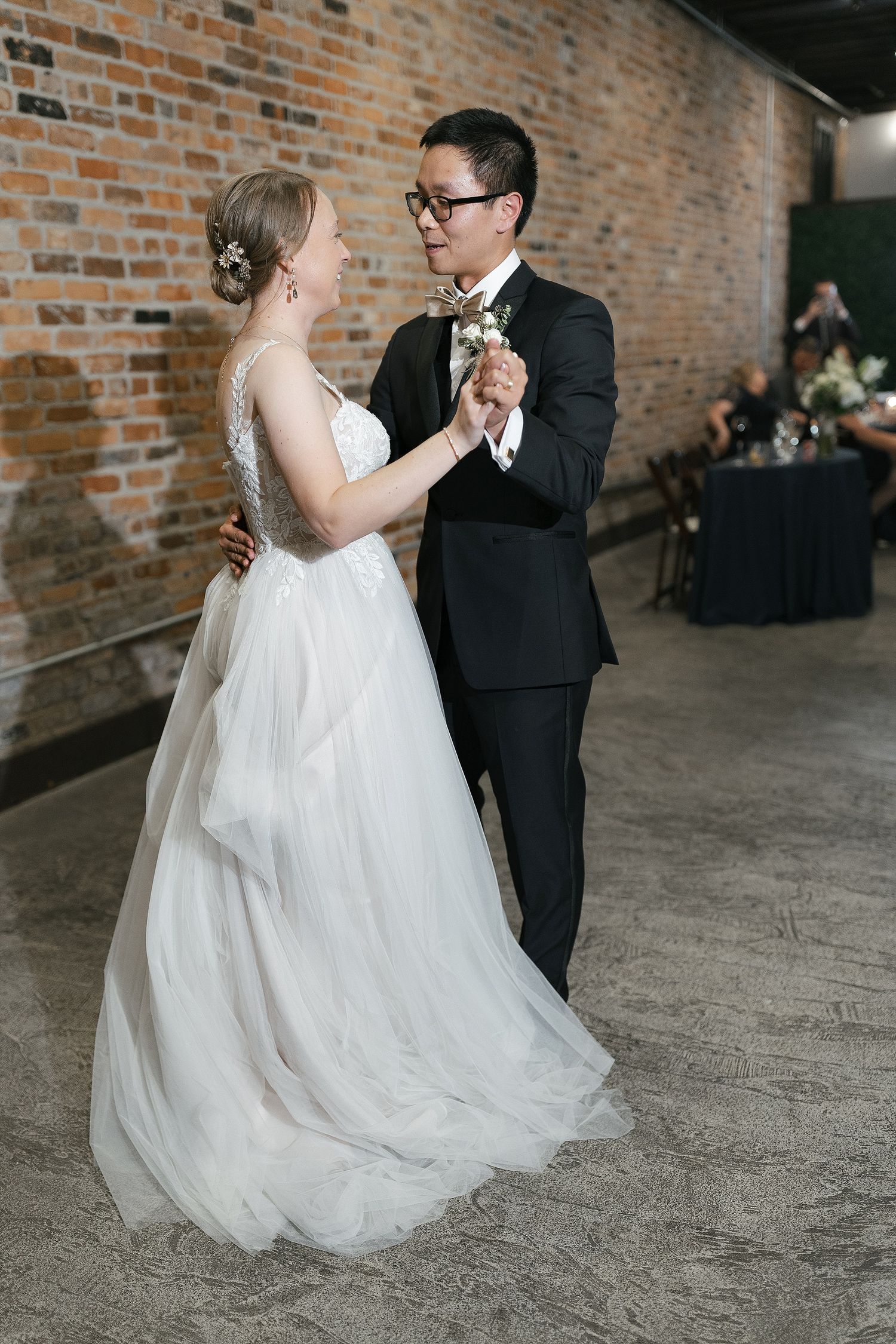 A couple in wedding attire dancing together in front of an exposed brick wall in an industrial-style venue.