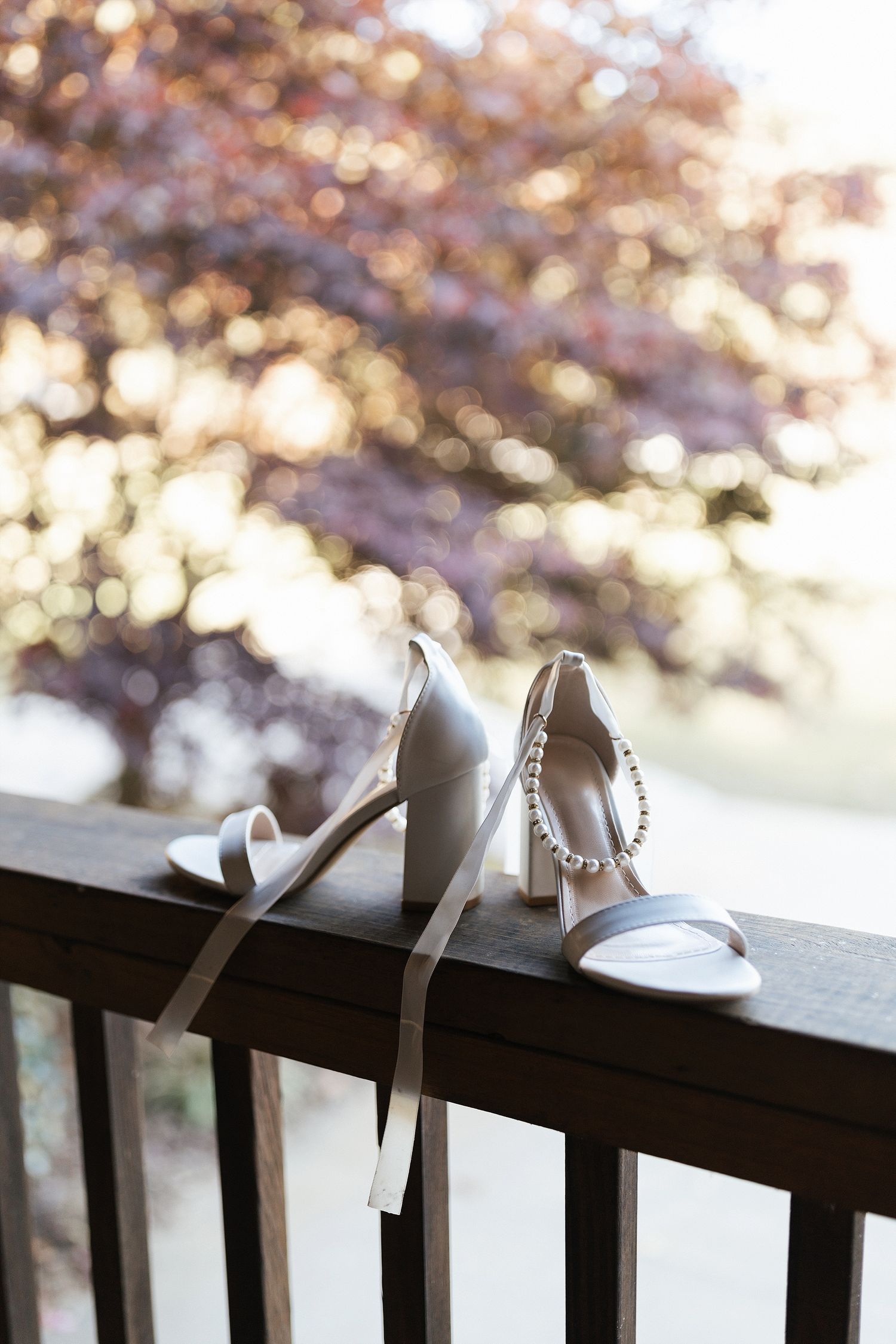 White bridal heels with sparkly straps rest on a dark wooden railing with soft bokeh background at sunset.