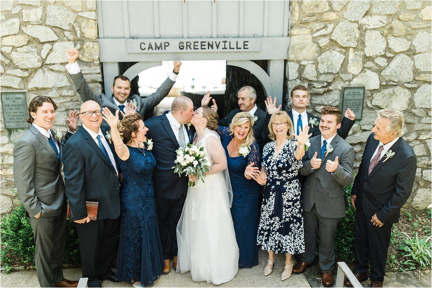 Wedding party poses in front of Camp Greenville stone entrance wearing navy and gray attire.
